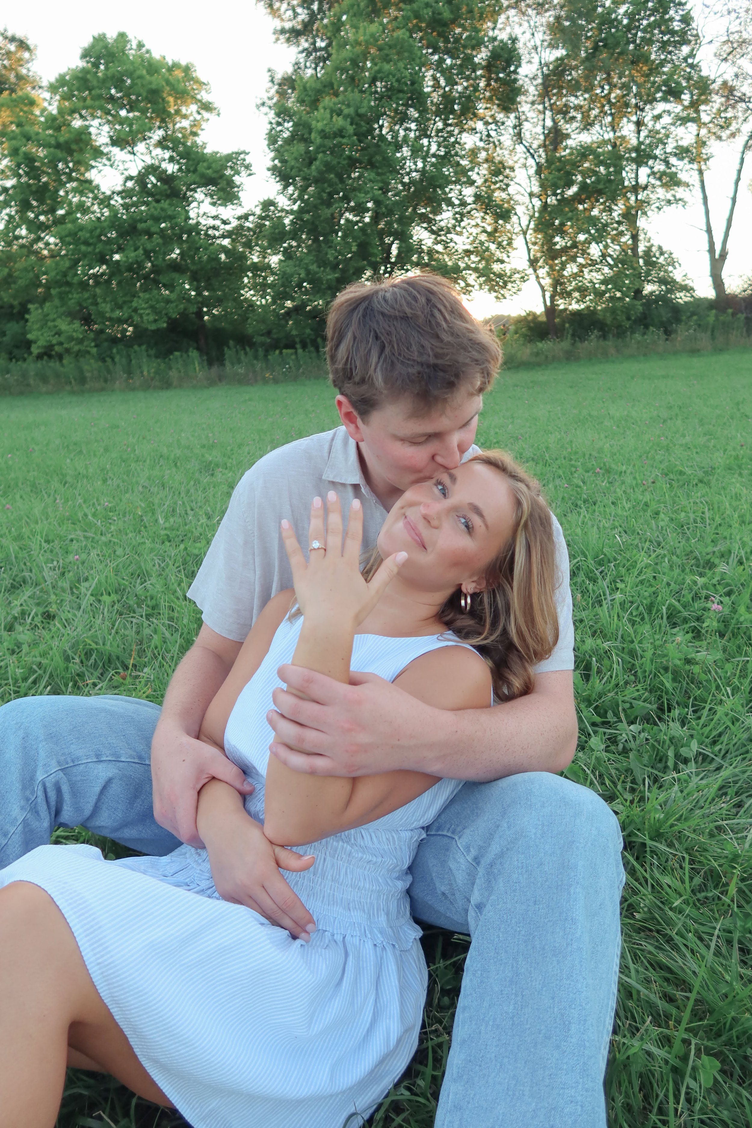 A young couple sitting on grass in a park, embracing and smiling. The man is kissing the woman on her forehead, and she is looking at the camera with a happy expression. The background features green trees and a clear sky.