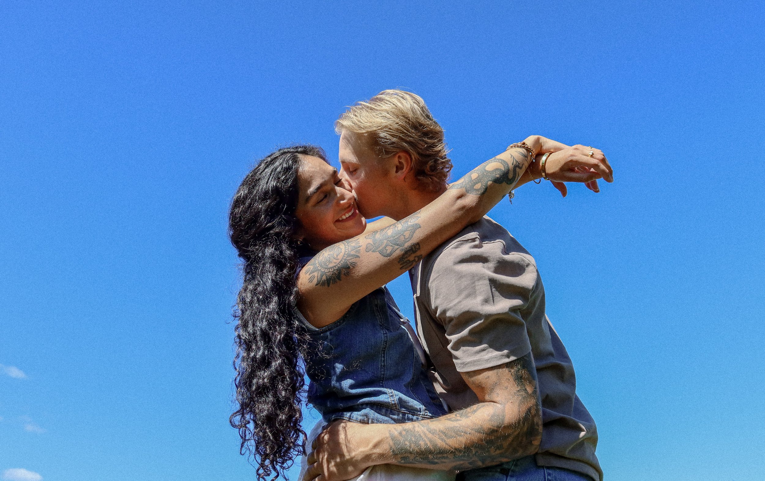 A couple embracing outdoors under a clear blue sky, with the woman smiling and the man kissing her cheek.