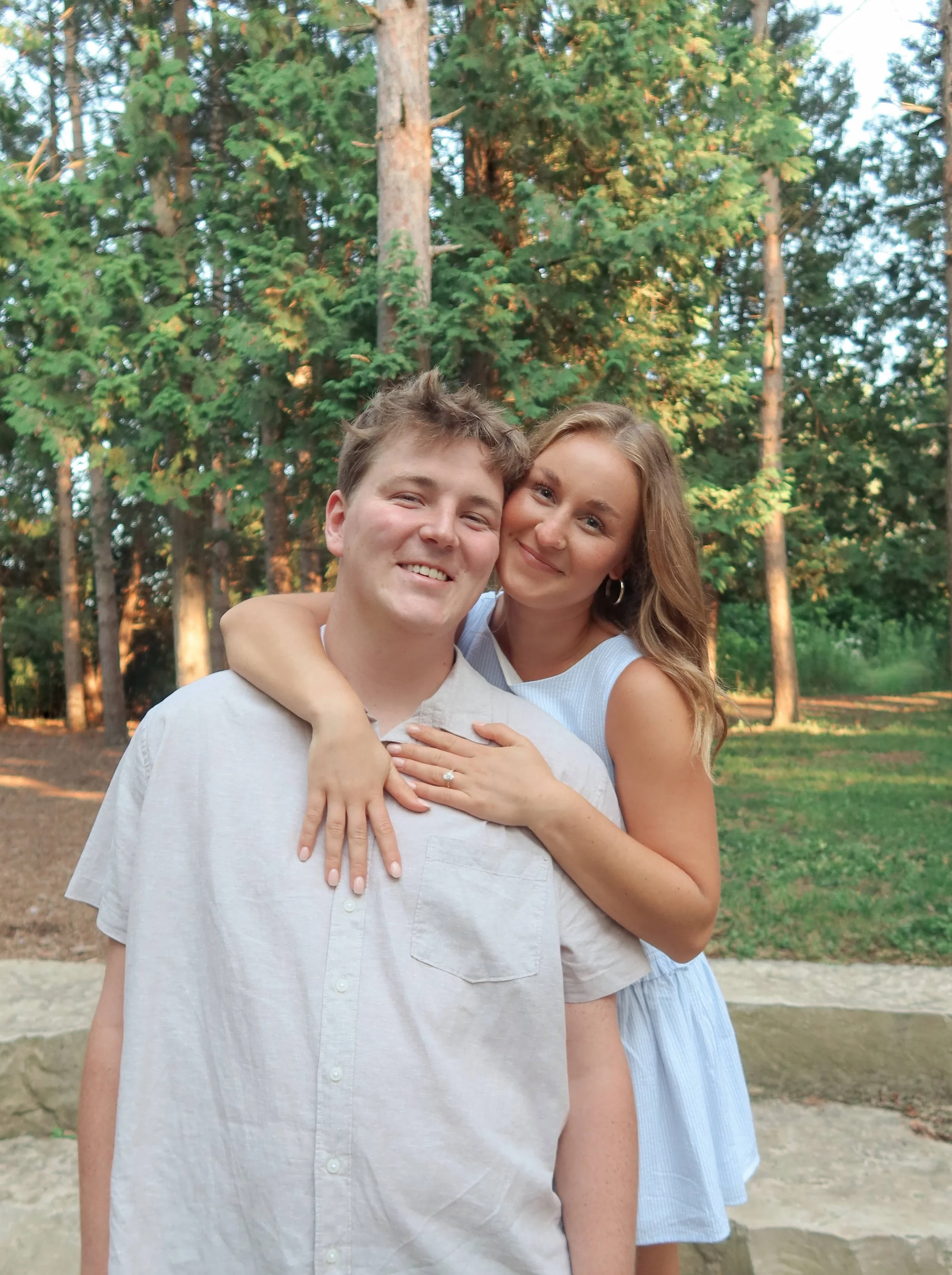 A smiling young man and woman embracing outdoors in front of a forest of tall green trees during the daytime.
