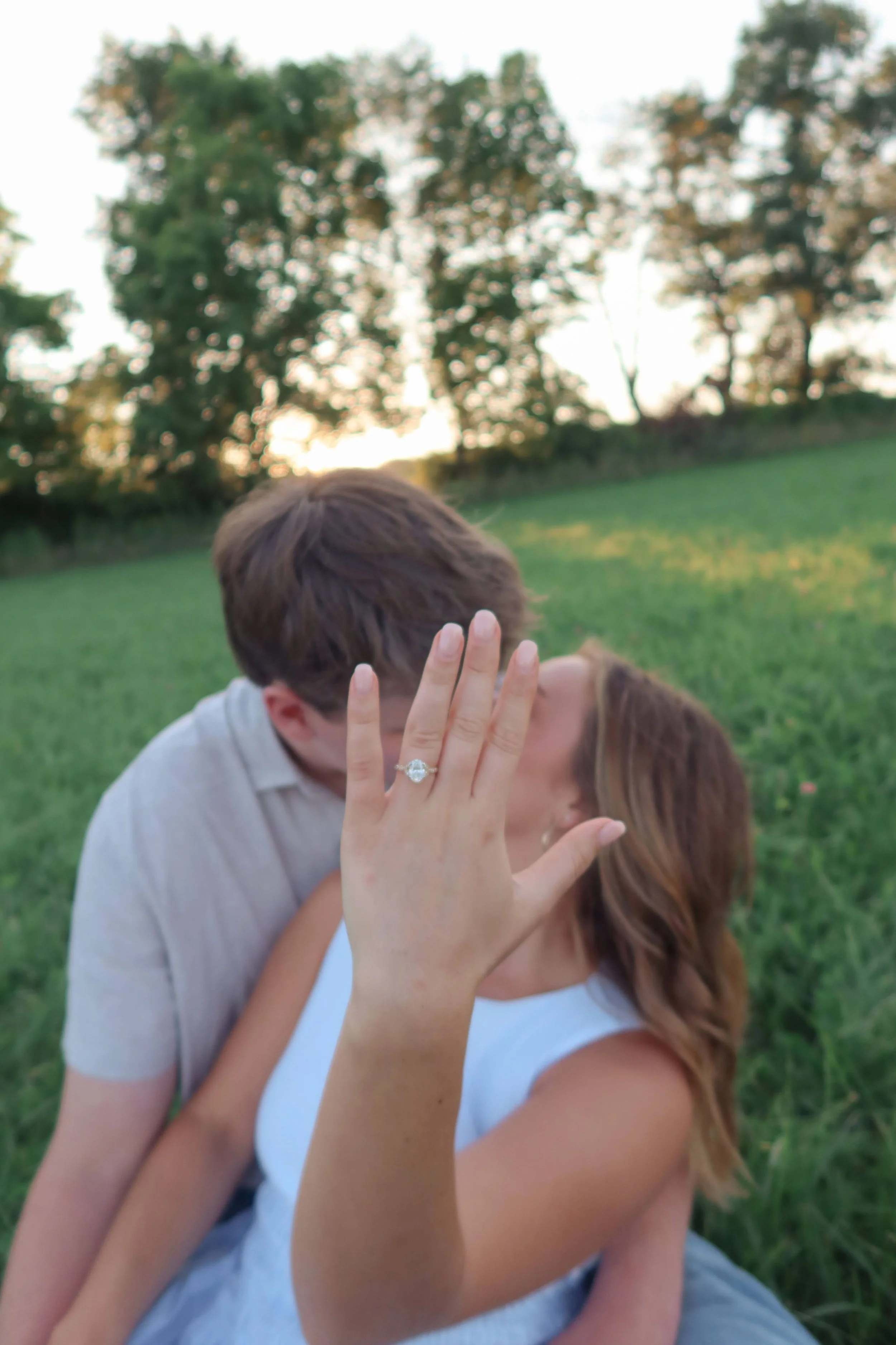 A couple is sitting on grass in a park during sunset, with trees in the background. The woman is showing a ring on her finger, indicating an engagement or wedding.