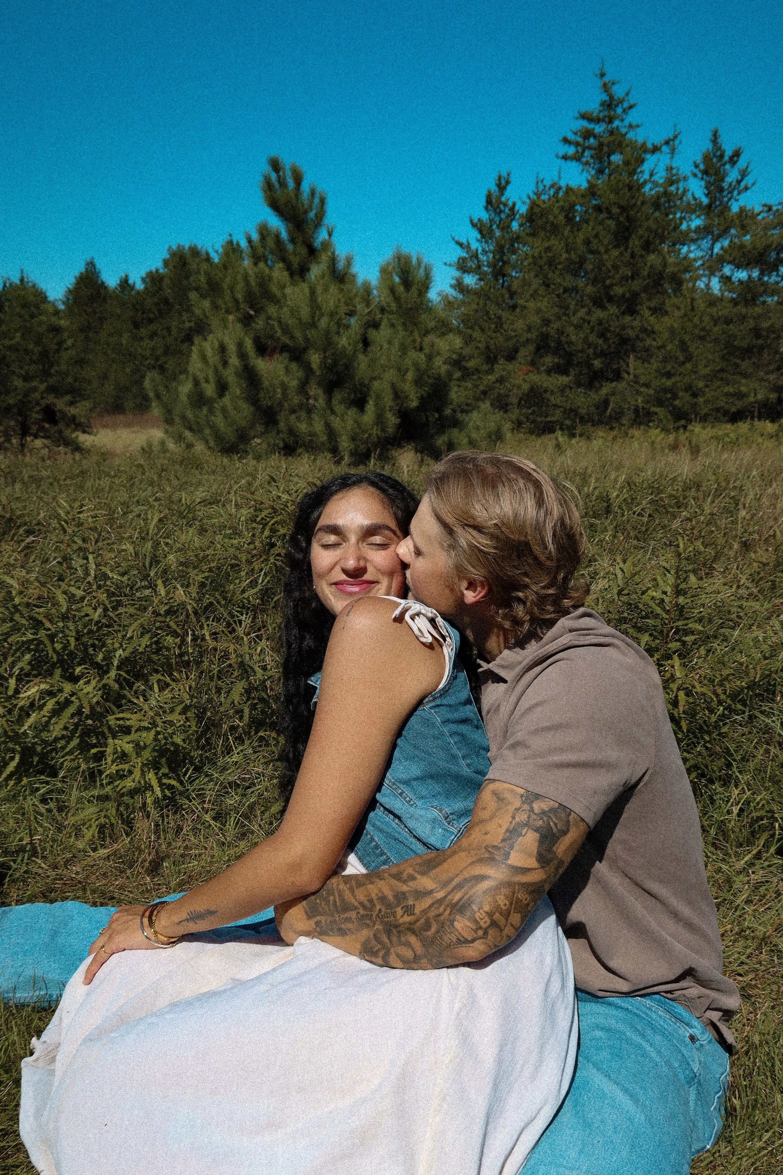 A couple sitting outdoors in a grassy field with trees and a blue sky in the background. The woman has long dark hair and is smiling with her eyes closed, while the man with short light brown hair is kissing her cheek.