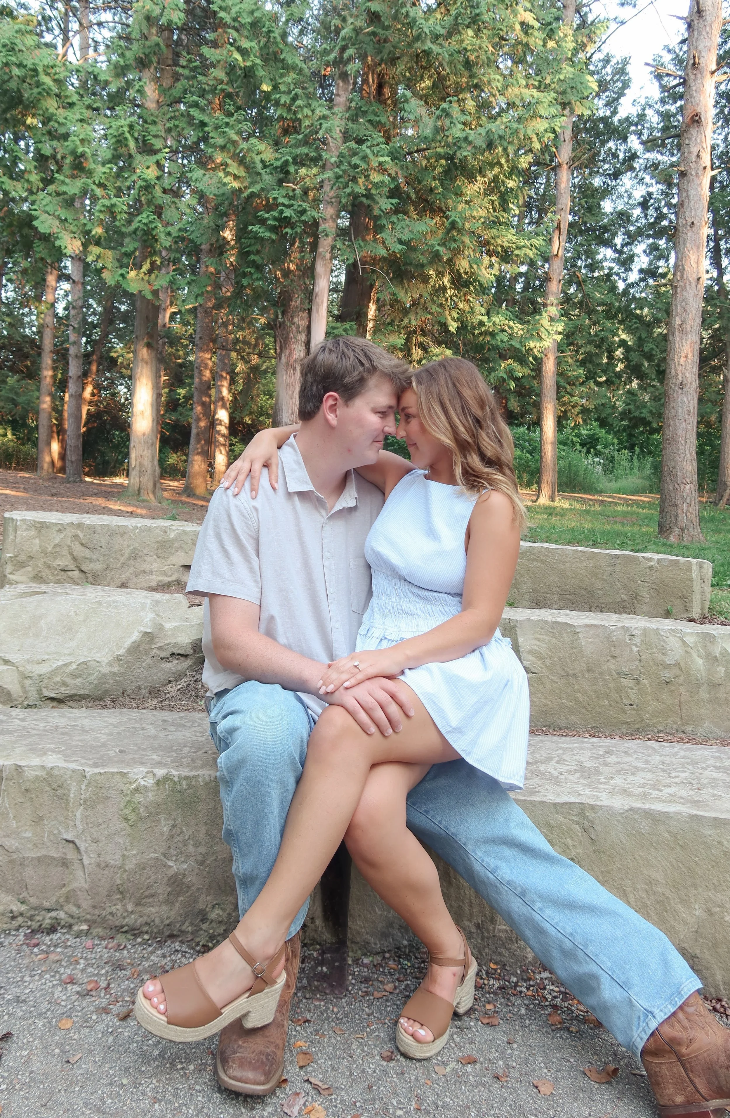 A couple sitting on stone steps outdoors, with trees in the background, engaging in a close, affectionate pose with foreheads touching.