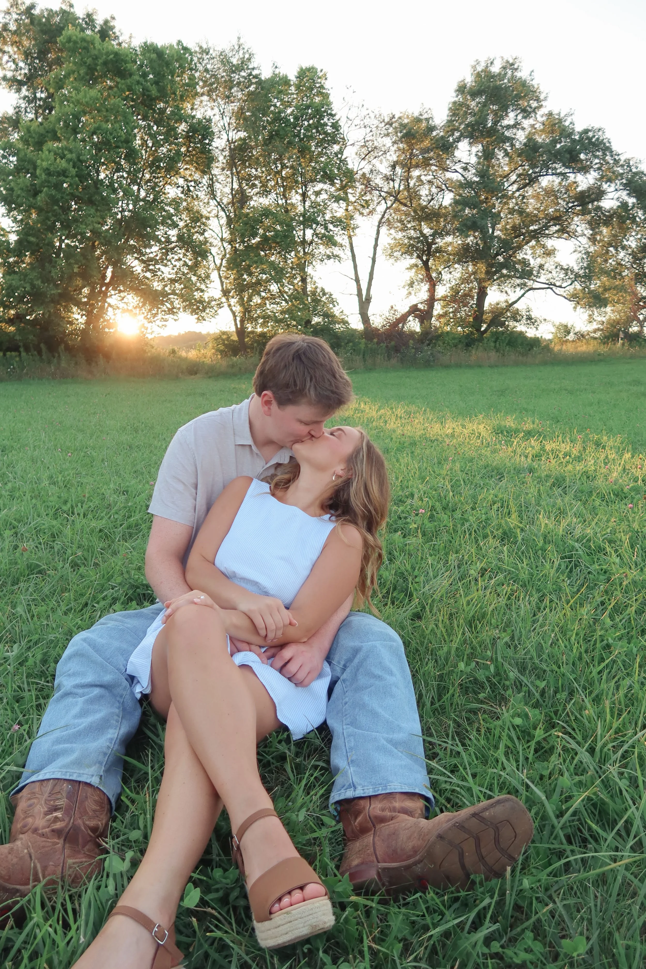 A young couple sitting on grass in a field, kissing, with the sun setting behind trees in the background.