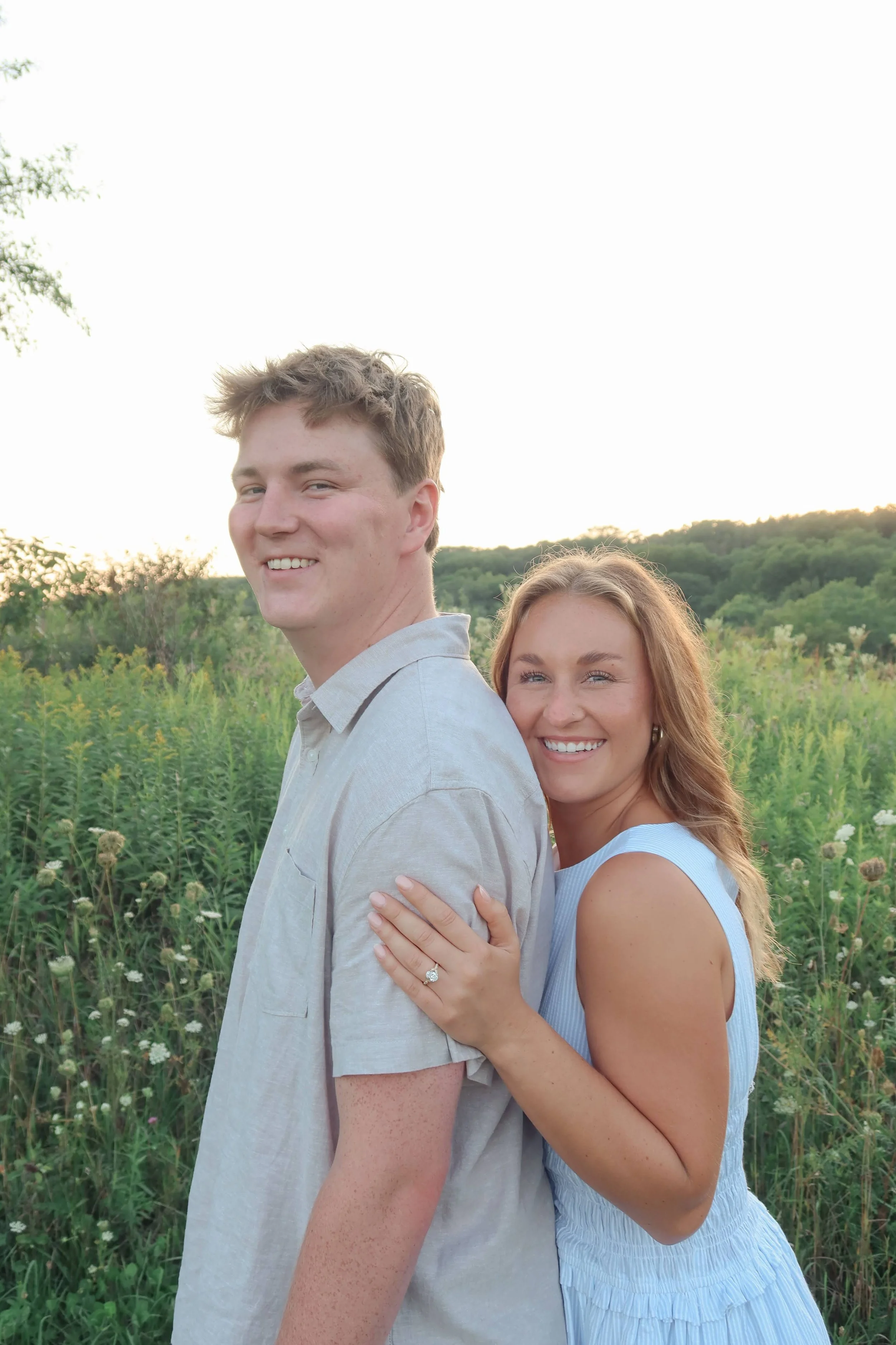 A smiling young woman with light brown hair and blue eyes hugging a smiling young man with light brown hair and blue eyes from behind in a field of green plants during sunset.