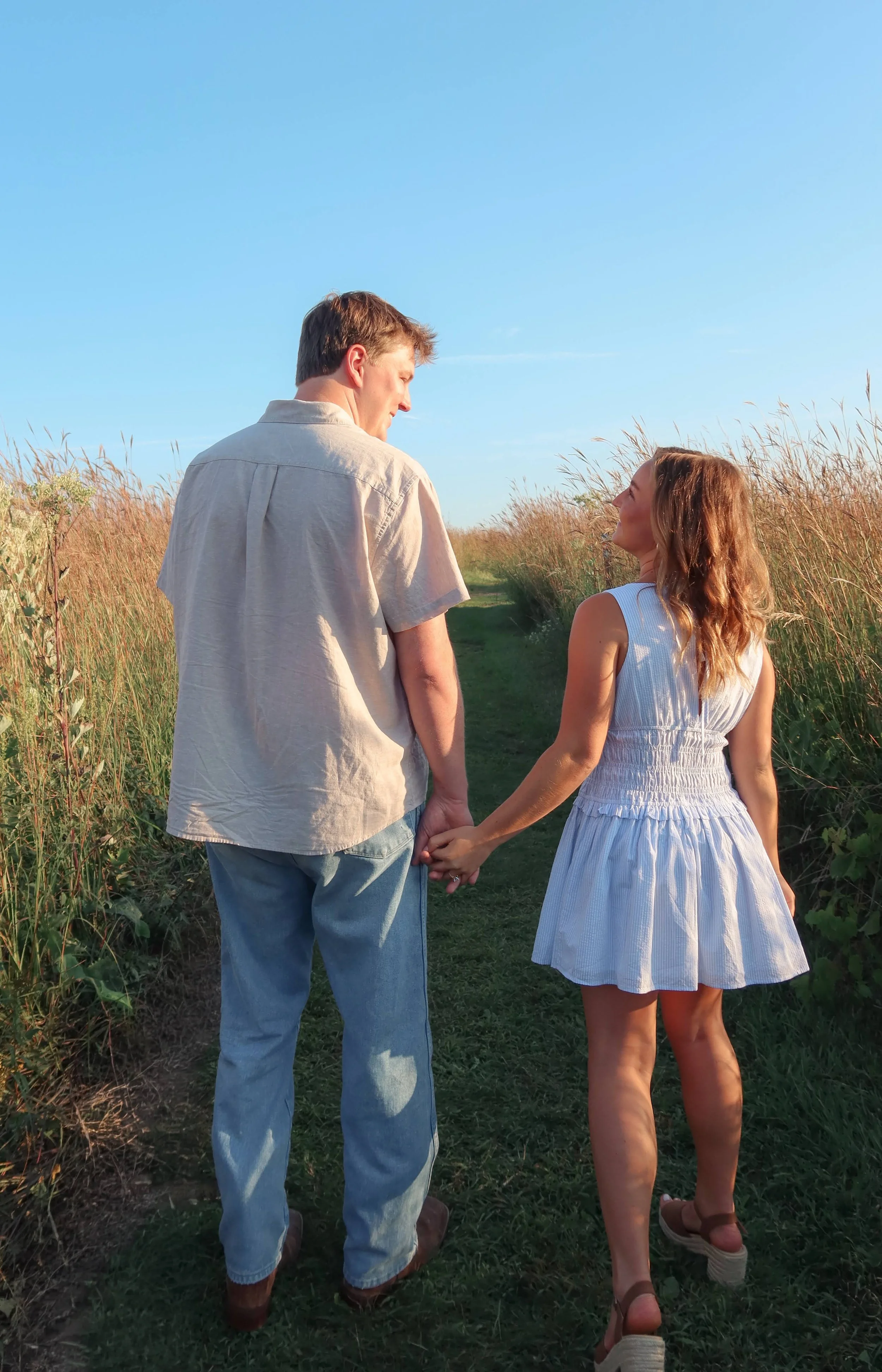 A young man and woman holding hands walking through a grassy trail in a field, facing each other and smiling under a clear blue sky.