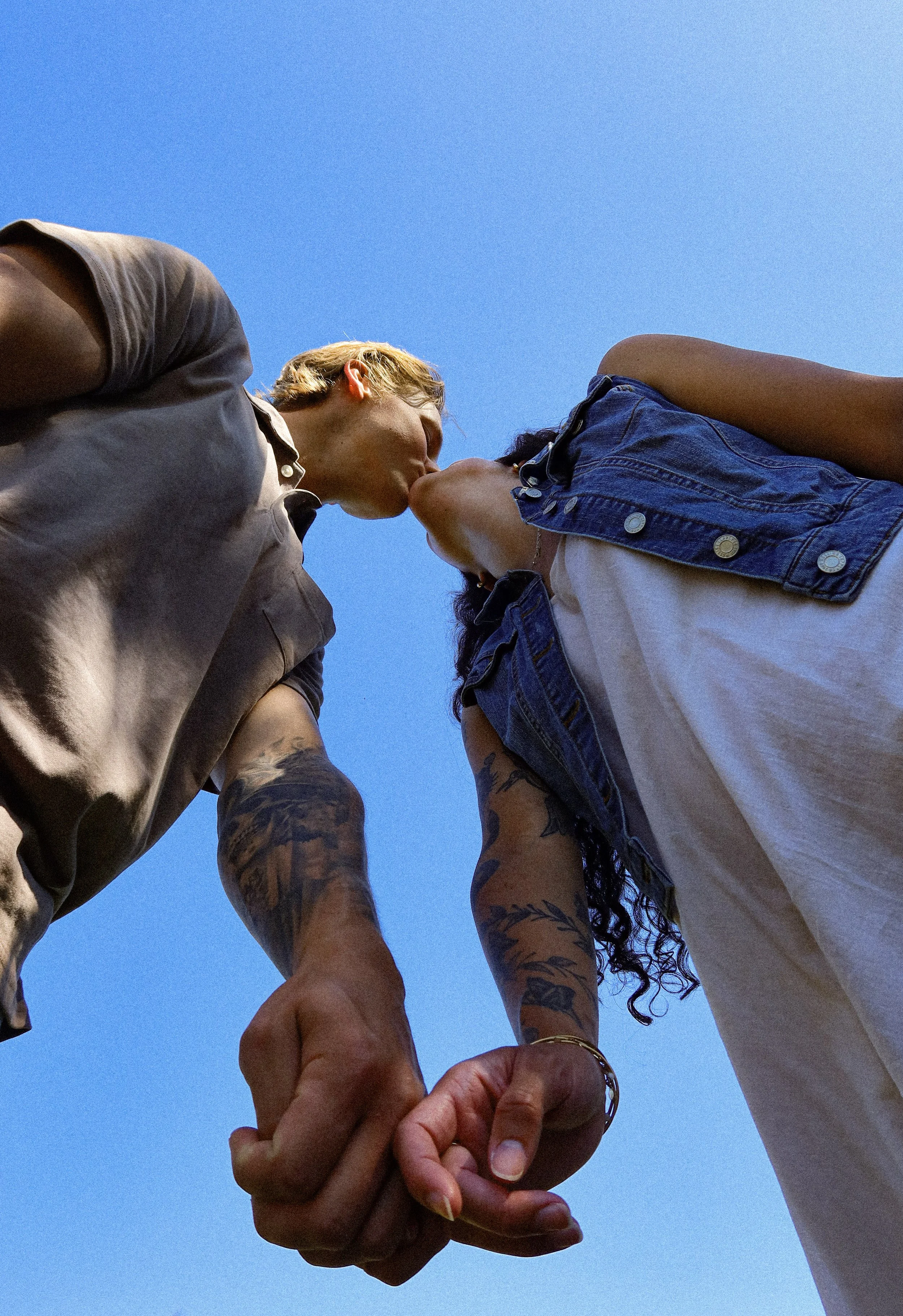 A couple holding hands and sharing a kiss outdoors against a clear blue sky, with visible tattoos on their arms.