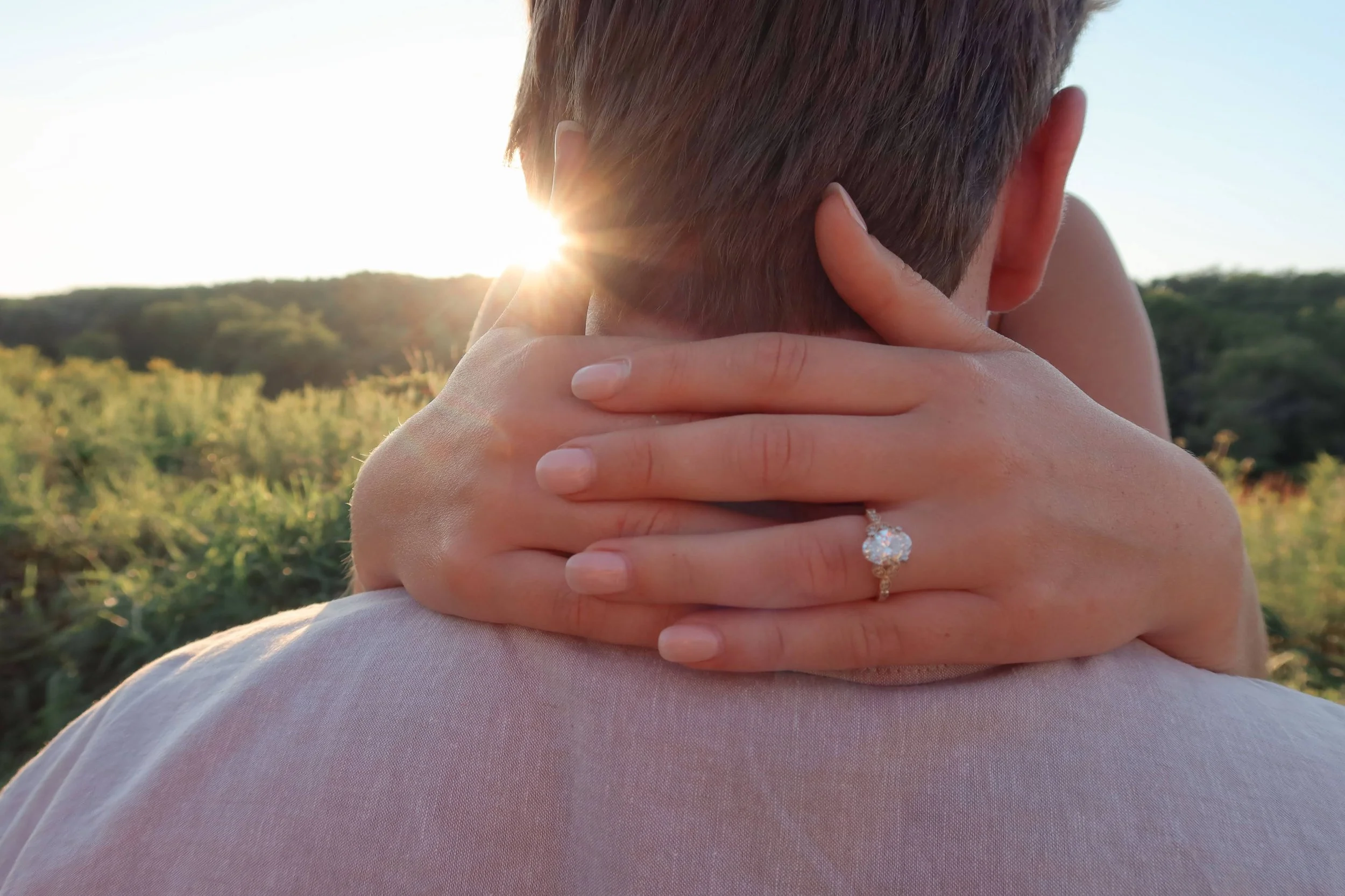 Close-up of a couple embracing outdoors with the setting sun in the background; focus on woman's hands holding man's head, showing an engagement ring.
