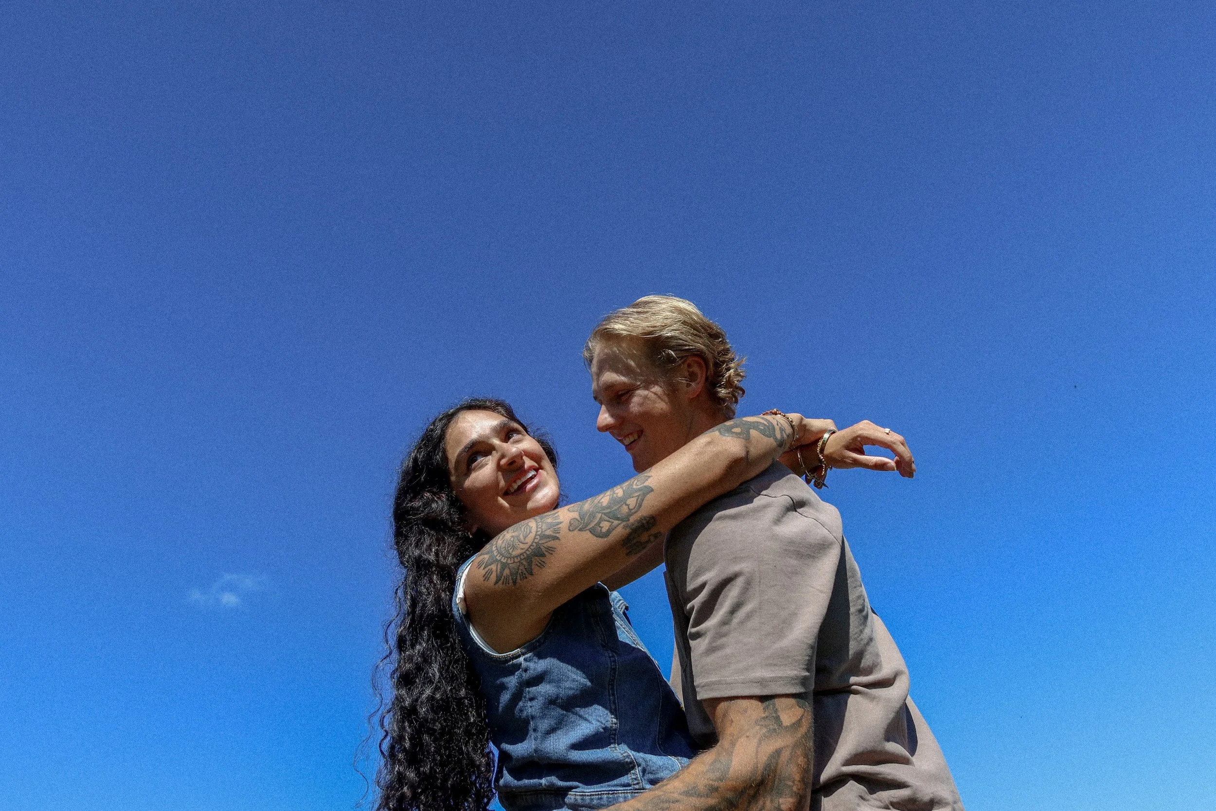 A man and woman are smiling and embracing each other against a bright blue sky.