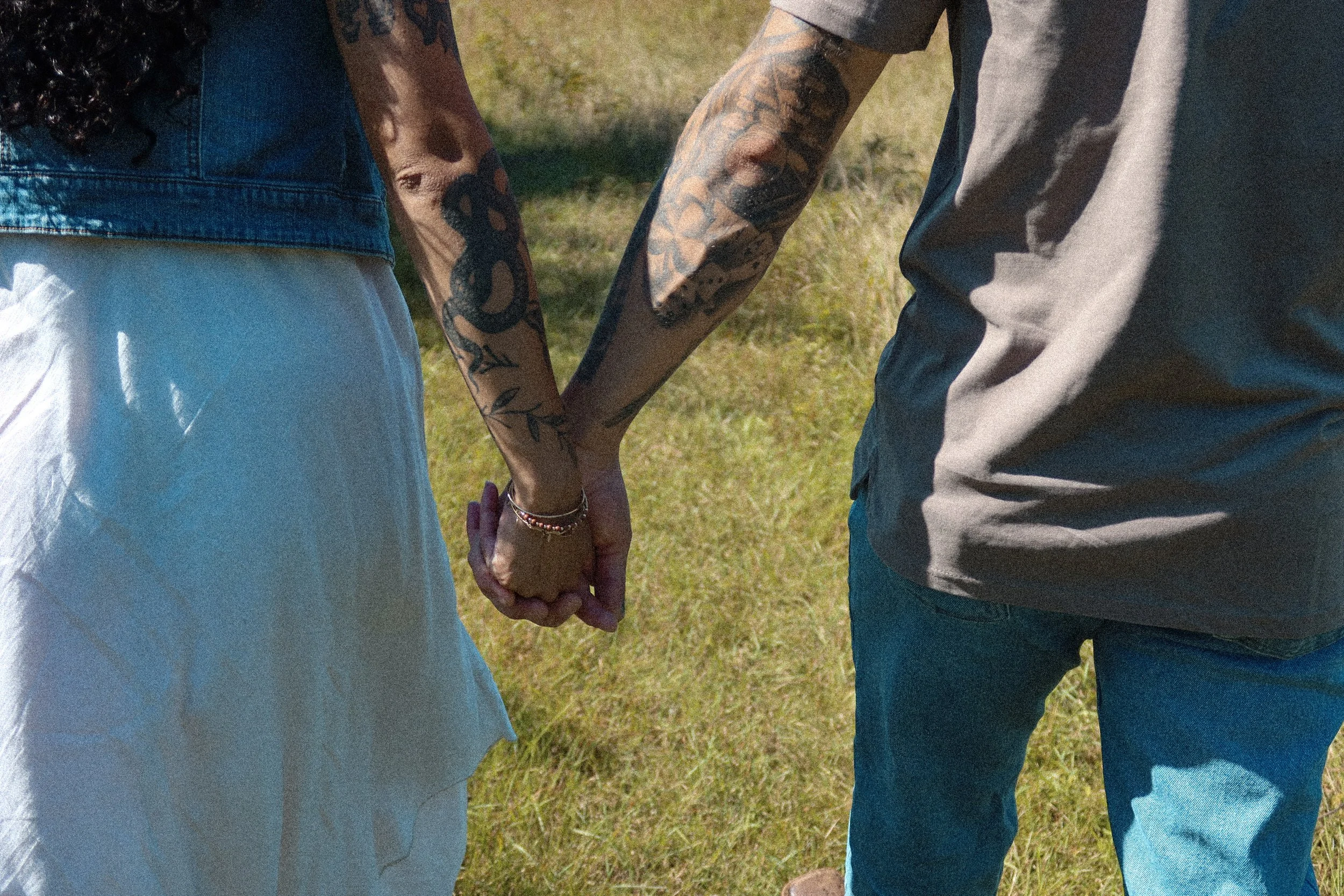 A close-up of a couple holding hands outdoors on grass, with the woman wearing a denim jacket and the man in a gray shirt. Both have tattoos on their arms.