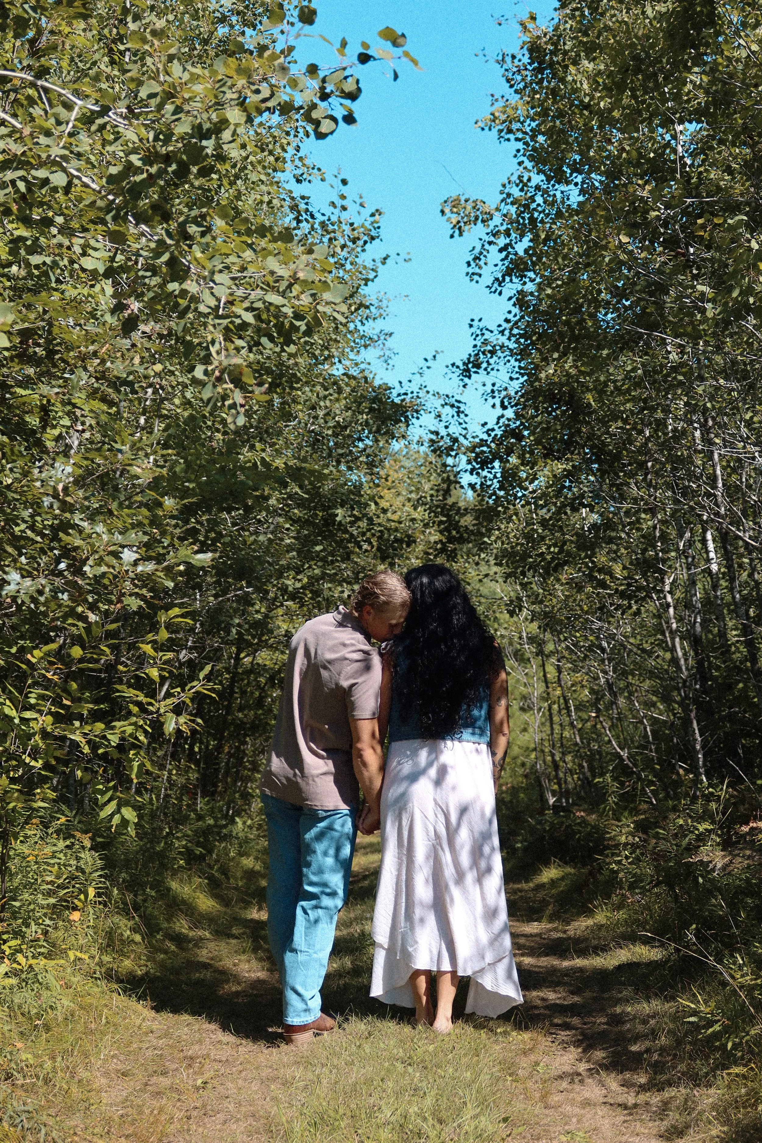 A couple holding hands and kissing in a forested area under a bright blue sky, surrounded by green trees and bushes.