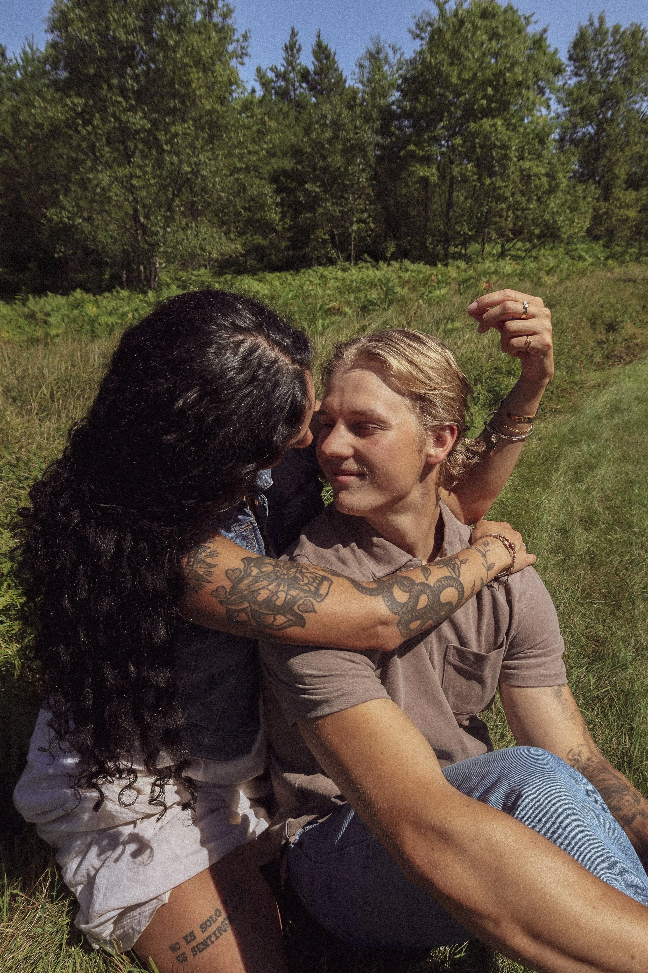 A woman with tattoos and a man with shaved blond hair sitting on grass outdoors, embracing closely with their foreheads touching, surrounded by green grass and trees under a clear sky.