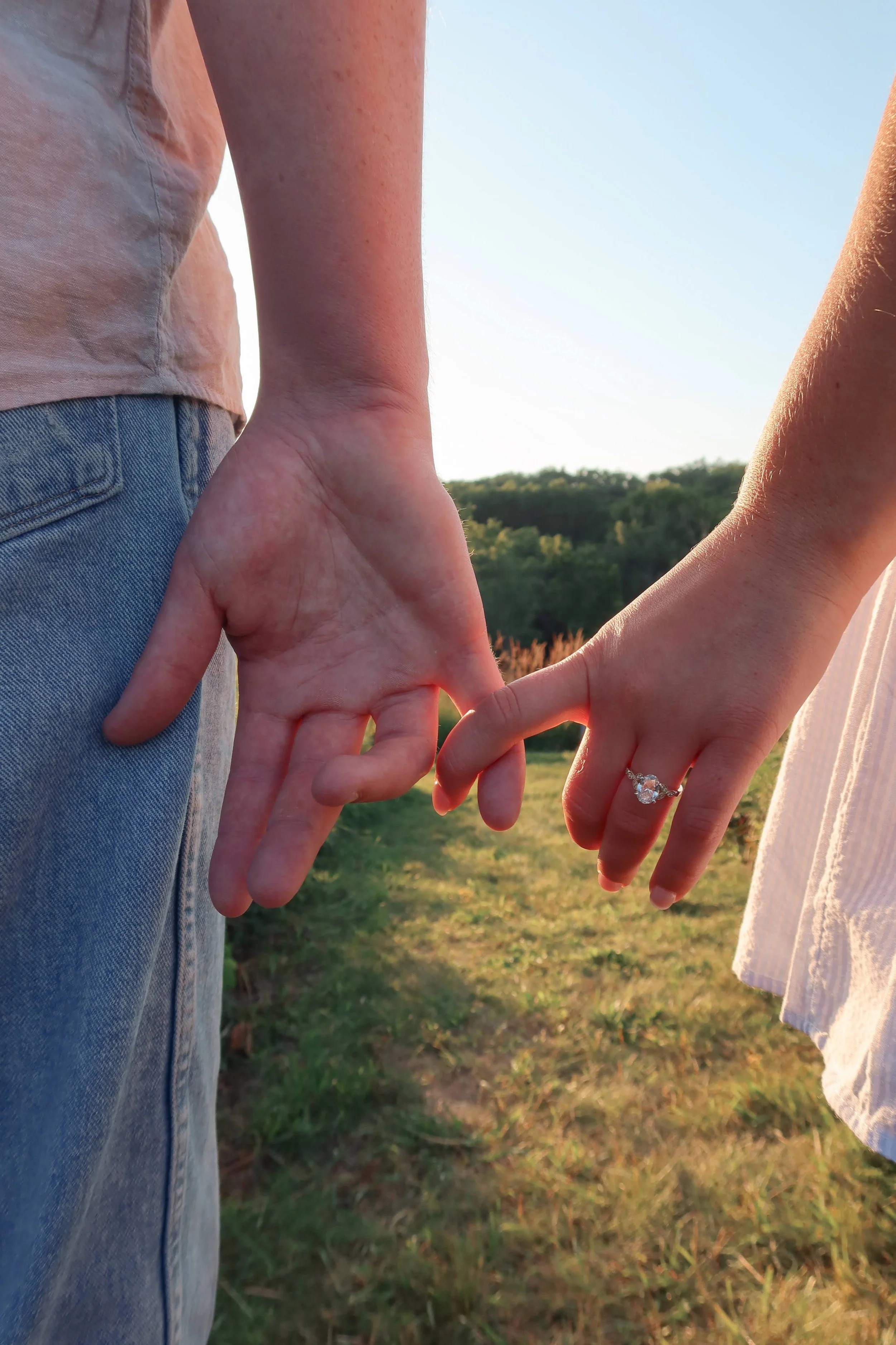 A close-up of a couple holding hands outdoors during sunset, with a background of green trees and a clear blue sky.