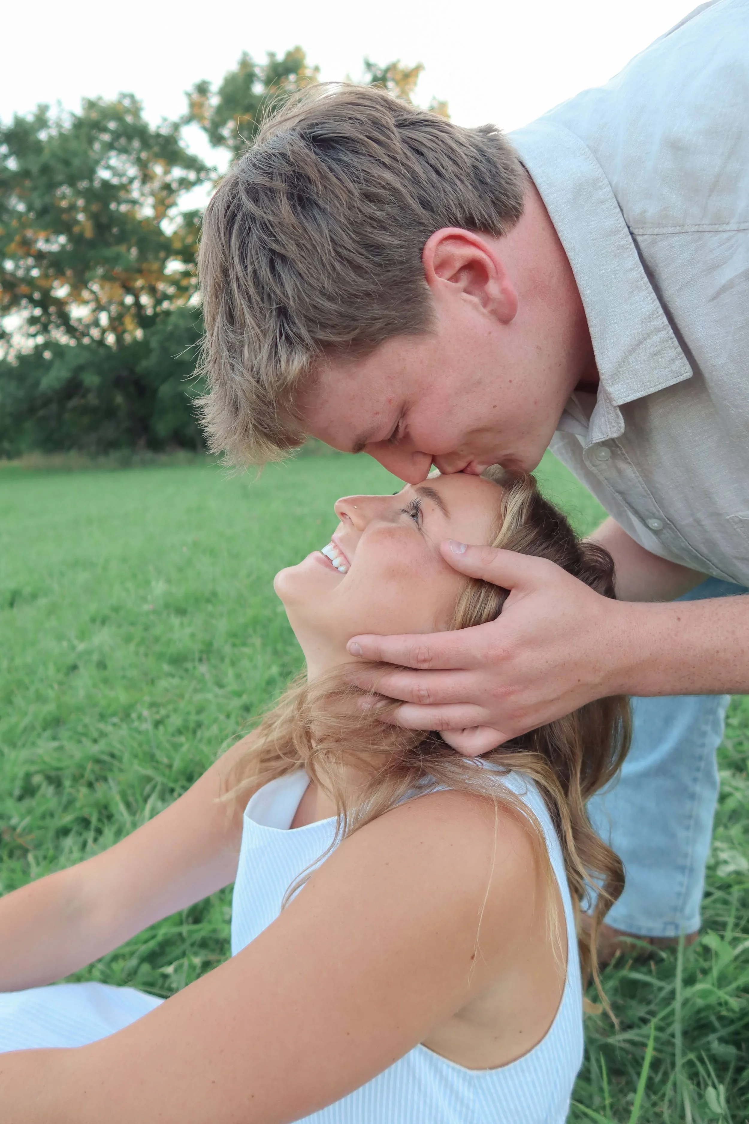 A young woman with long blonde hair lying on green grass, smiling and looking up at a man who is kissing her on the forehead. The man has short brown hair and is wearing a light-colored shirt. In the background, there are trees and a bright sky.