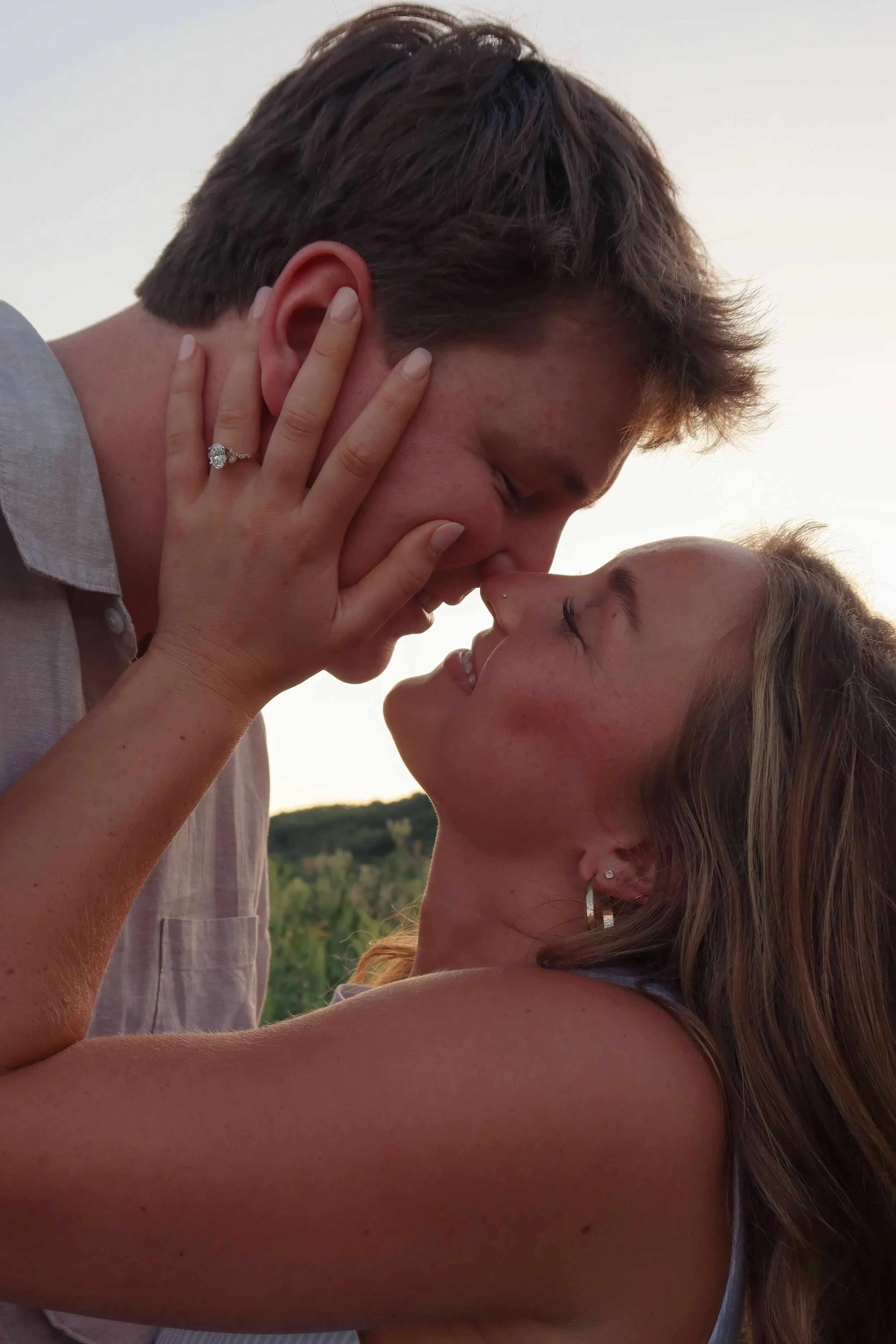 A couple is about to kiss, with the man gently holding the woman's face, their eyes closed and noses touching outdoors during sunset.