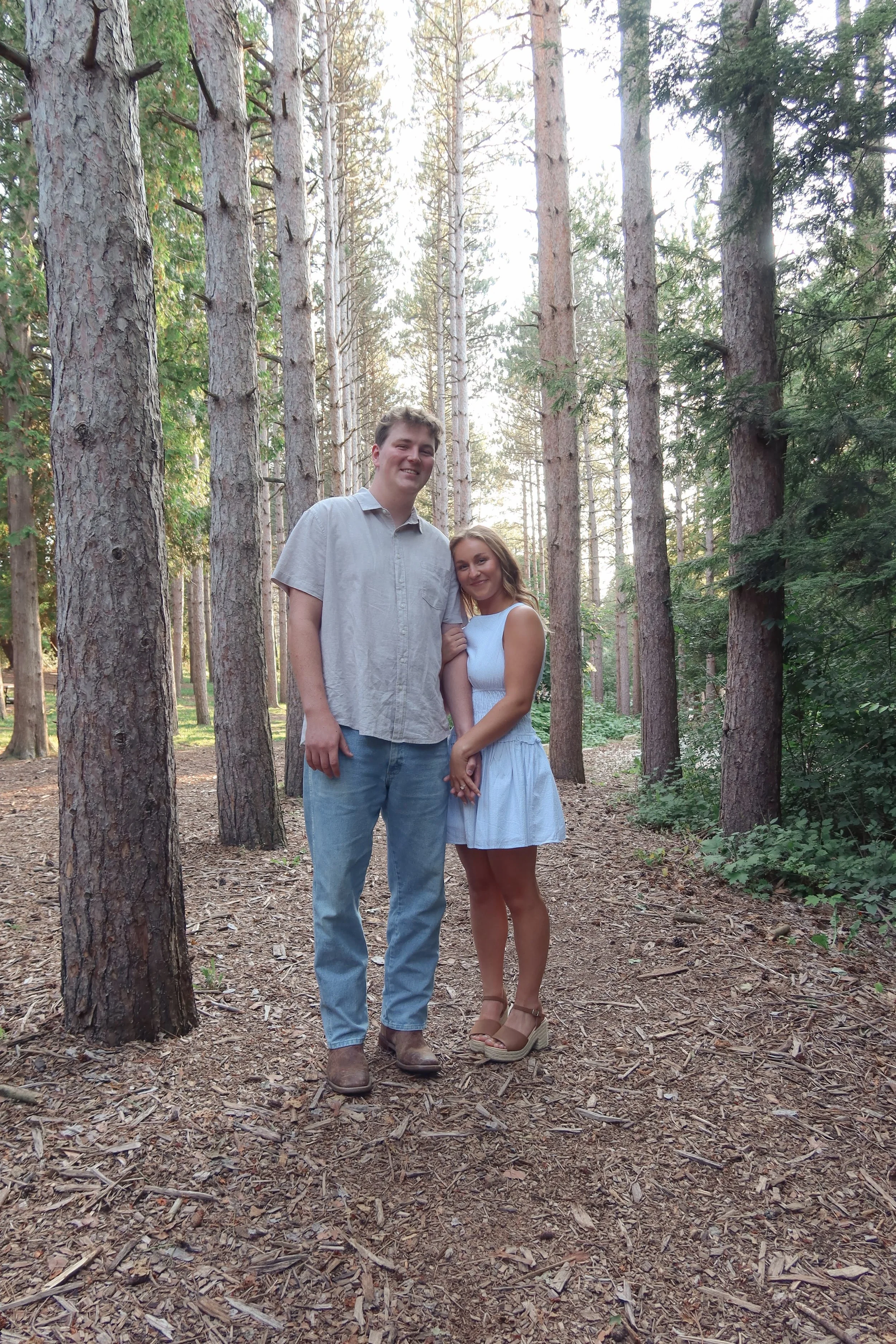 A young man and woman standing together in a forest. Tall pine trees surround them, and the sunlight filters through the branches. The man wears a light grey shirt and jeans with boots, and the woman wears a white dress with wedge sandals.