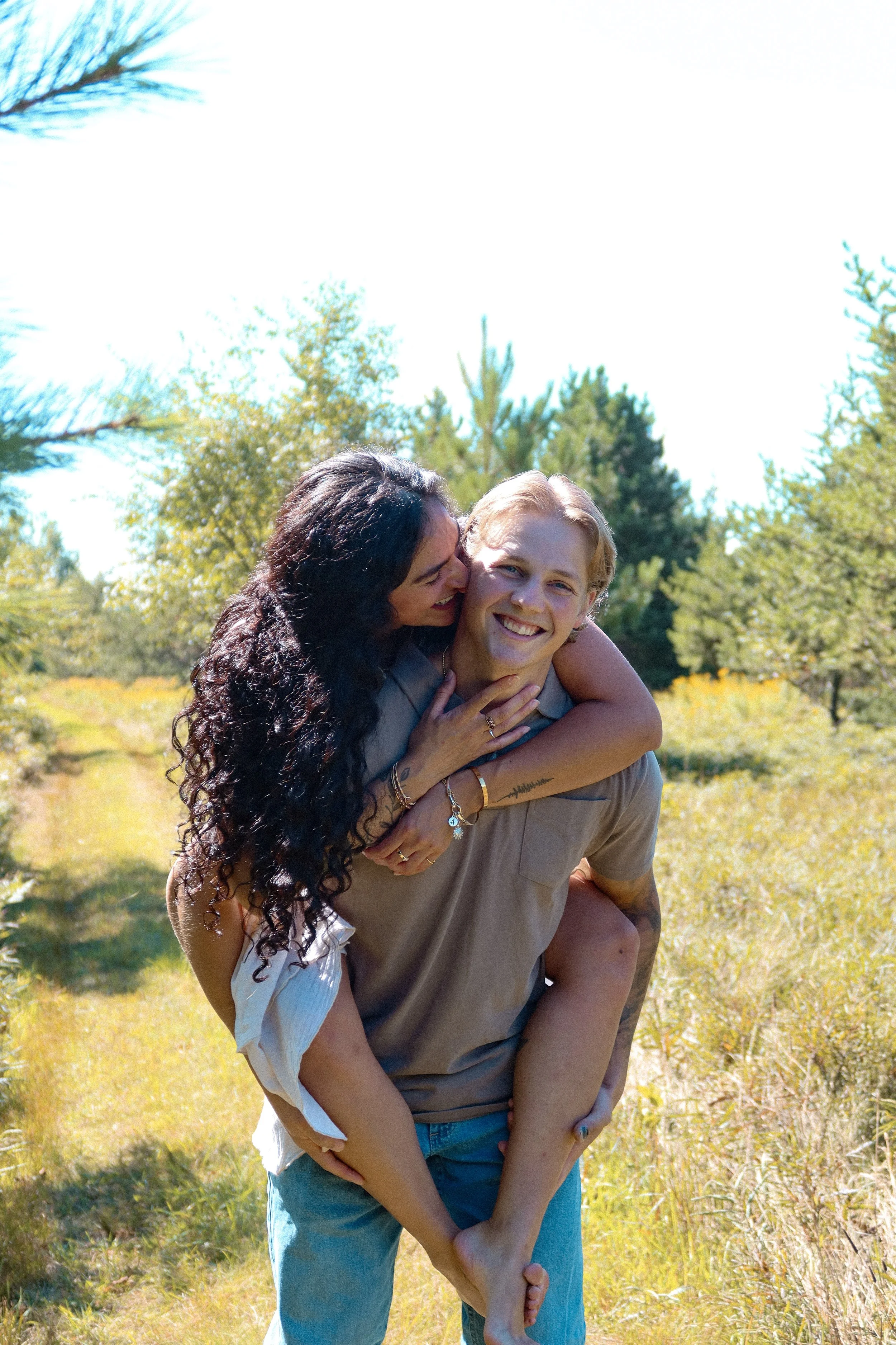 A young man carrying a young woman on his back in a sunny, green outdoor setting with trees and grass.