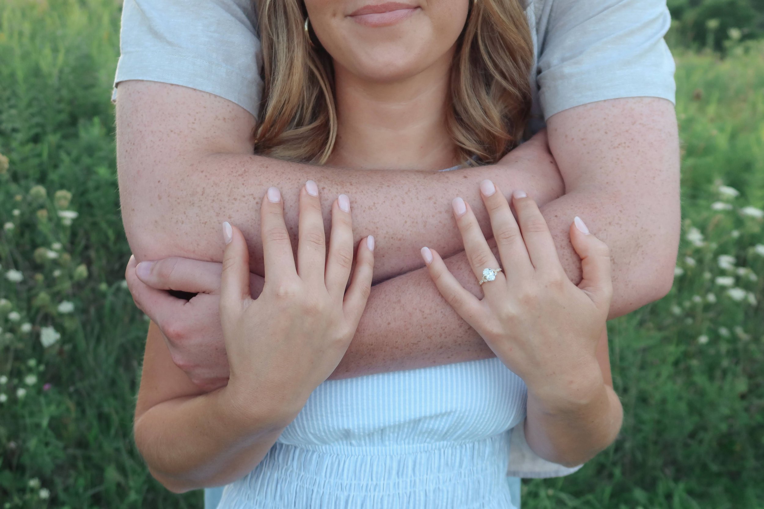 A woman with light skin and freckles is being embraced from behind by another woman. The woman in front has her arms crossed over her chest, showing a wedding ring on her left hand. The second woman's arms are wrapped around her. They are outdoors in