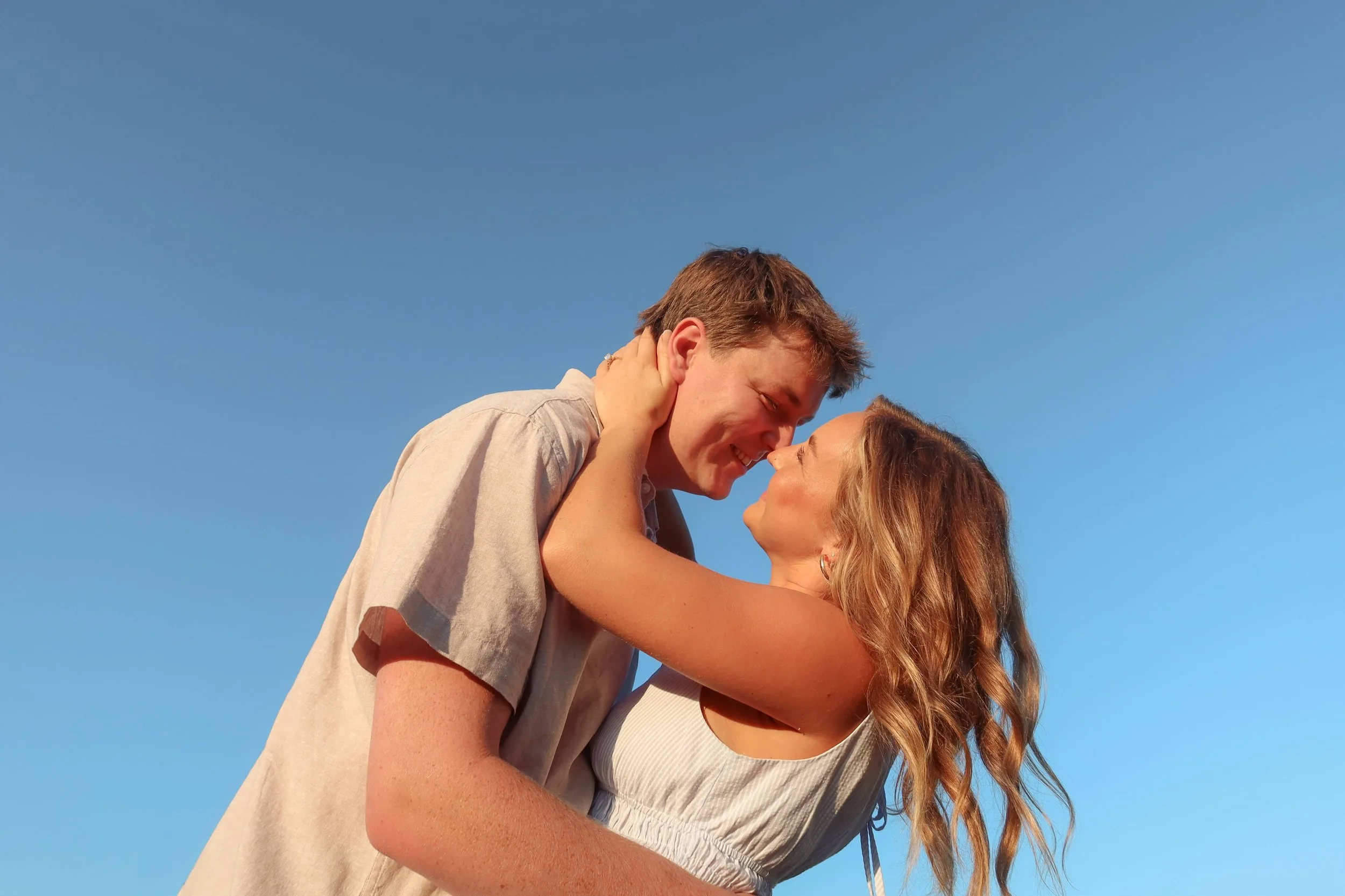 A young couple is embracing and smiling with their noses touching against a clear blue sky, sharing an intimate moment outdoors.