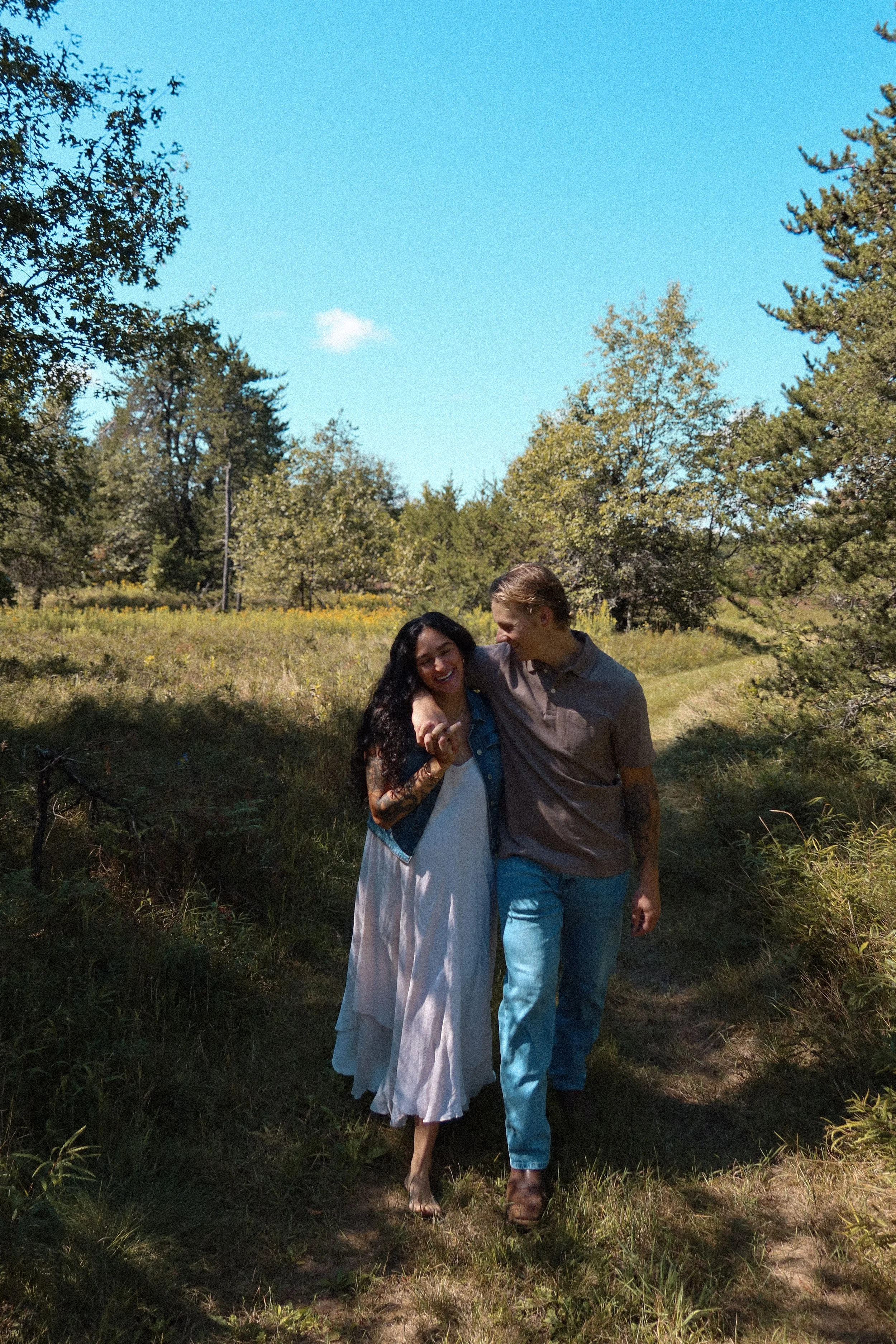 Two people, a woman with long dark hair wearing a white and blue dress, and a man with light brown hair wearing a gray shirt and blue jeans, are walking outdoors in a lush green area with trees and a bright blue sky, smiling and laughing together.
