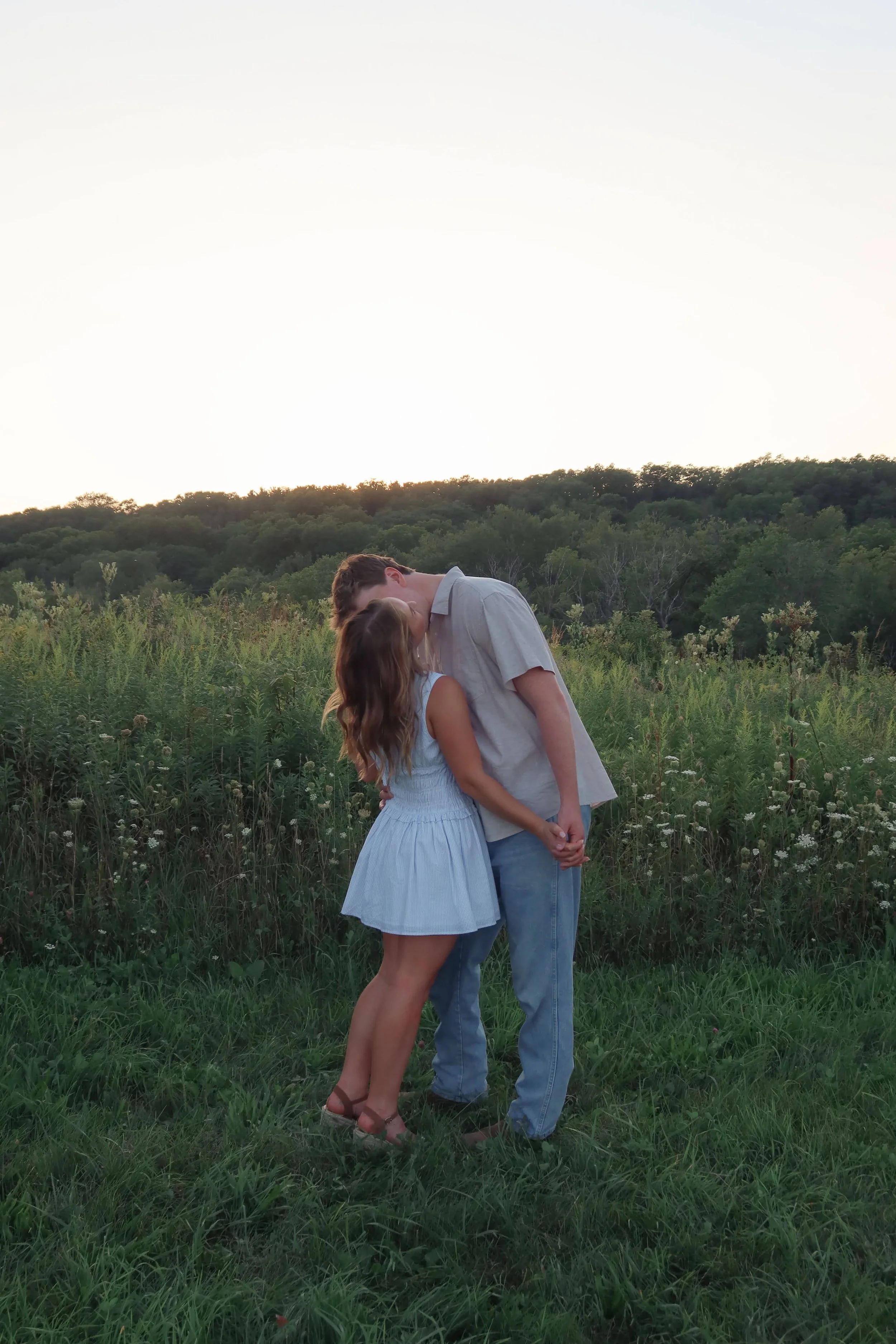 A couple sharing a kiss in a grassy field with green trees and hills in the background at sunset.