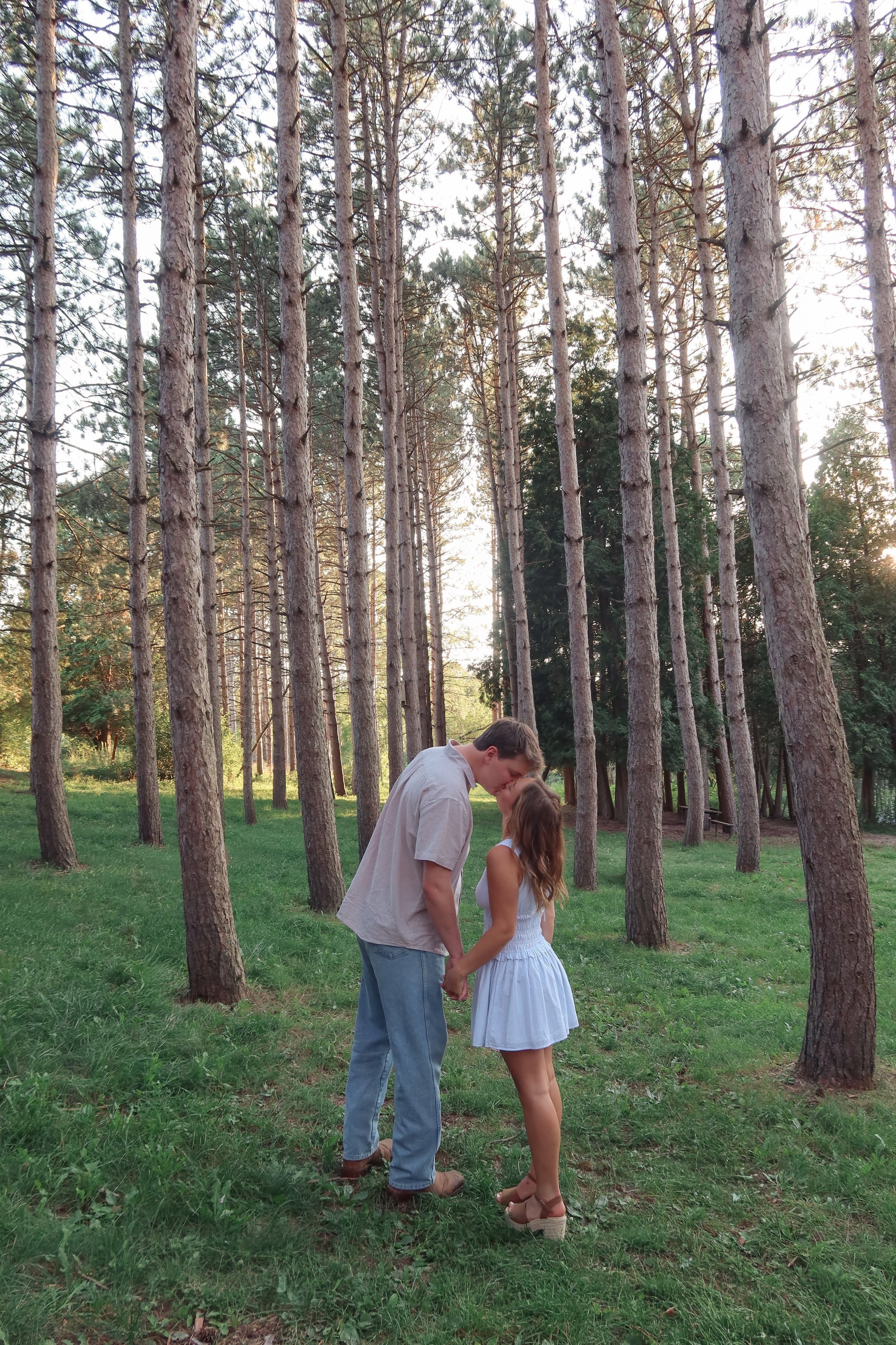 A young man and woman standing together in a forest, holding hands, with tall trees and greenery in the background, during sunset.