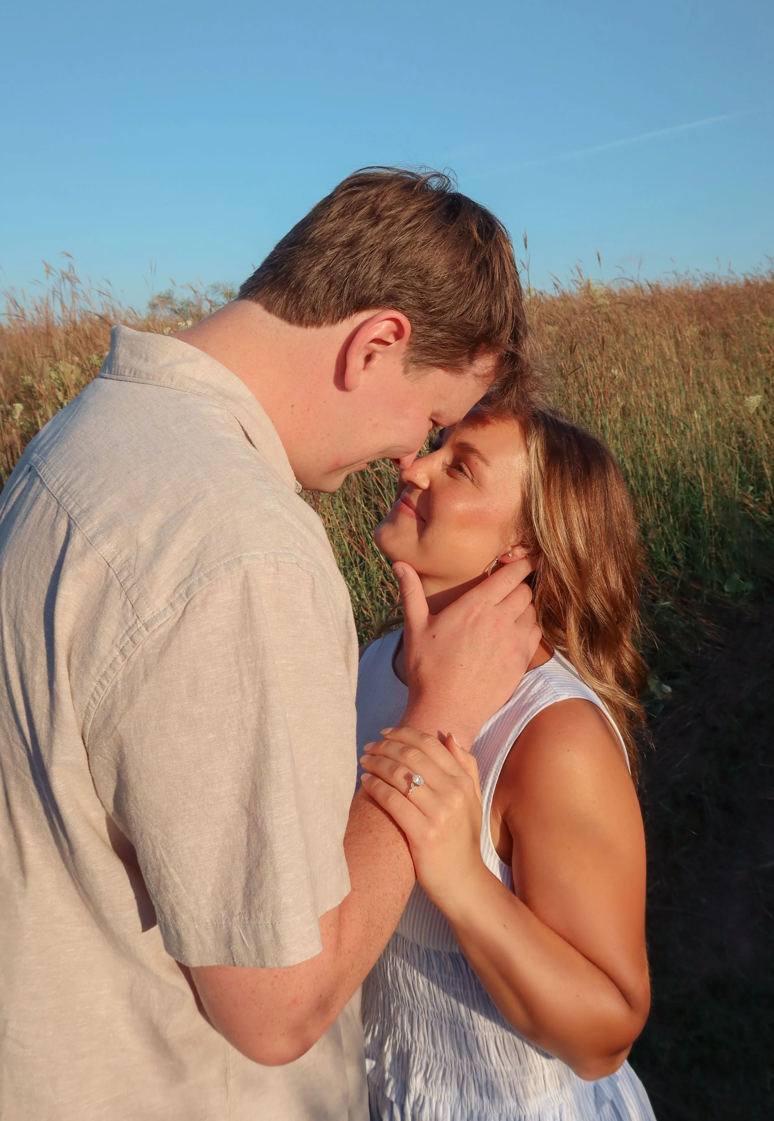 A couple is sharing a close moment outdoors, with the man leaning down towards the woman, who is looking up at him. The woman has long brunette hair and is wearing a white sleeveless dress and a ring. The background shows a grassy field under a blue sky.