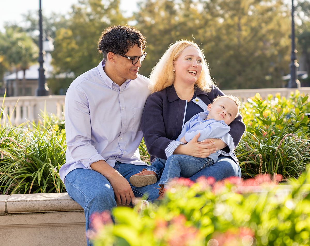 A family of three sitting outdoors on a sunny day, with a man, woman, and a young child. The woman is holding the child on her lap, all smiling and enjoying the moment. Greenery and trees are in the background.