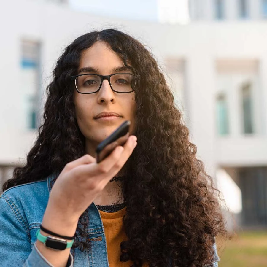 A young woman with long curly hair, glasses, and a denim jacket looking at the camera, holding a smartphone outdoors with a modern building in the background.
