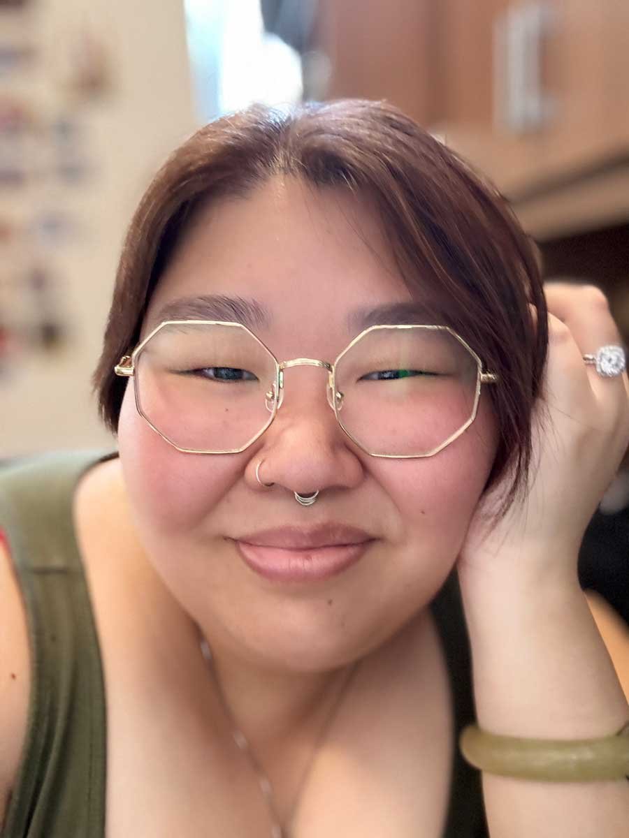 Close-up of a person with short brown hair, wearing large, geometric glasses, a nose ring, and a ring on their finger, smiling and resting their head on their hand.