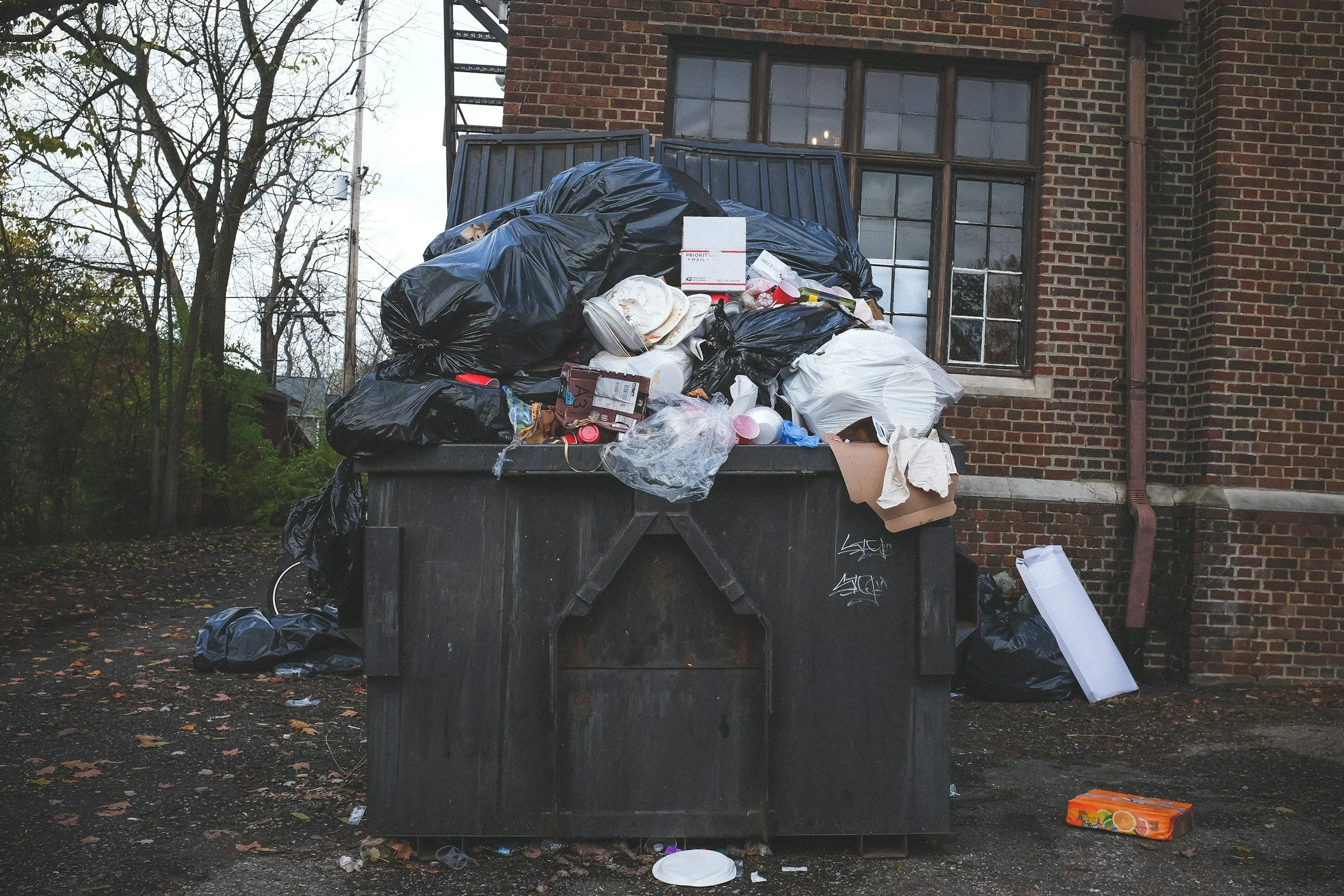 A large overflowing dumpster filled with black trash bags, paper plates, a pizza box, and discarded household items, situated outside a brick building with a multi-pane window, with trash and debris scattered on the ground around it.