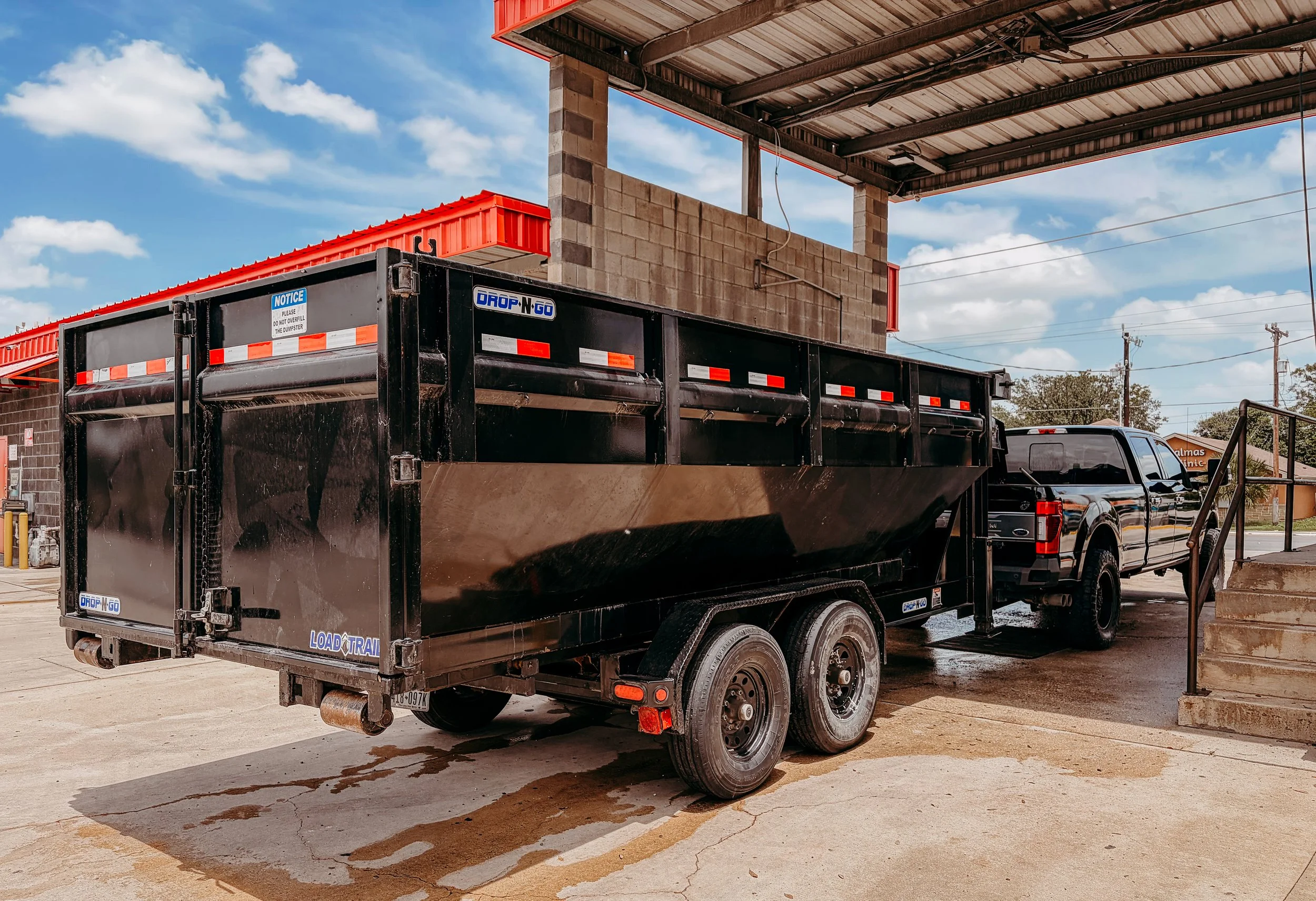 A black dump truck parked under a metal awning at a commercial area, with a boarded-up brick wall in the background and power lines above.