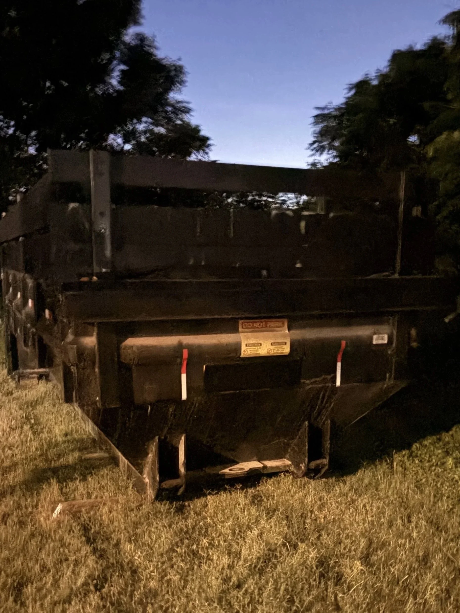 A trailer or container parked on grass with trees and a darkening sky in the background during dusk or dawn.