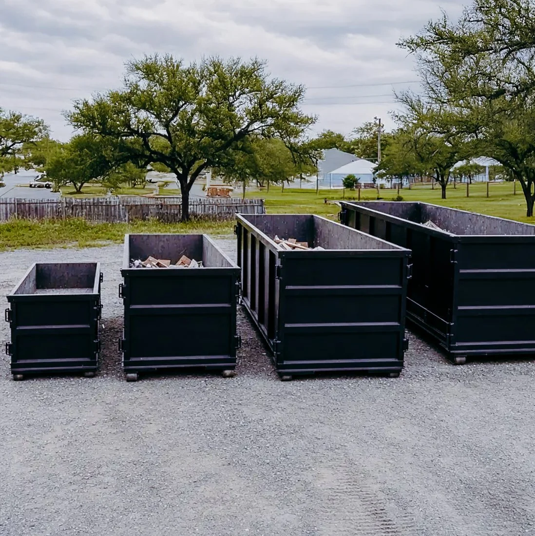 Four black dumpsters filled with trash sit on a gravel surface outdoors, with trees, grass, and buildings in the background under a cloudy sky.