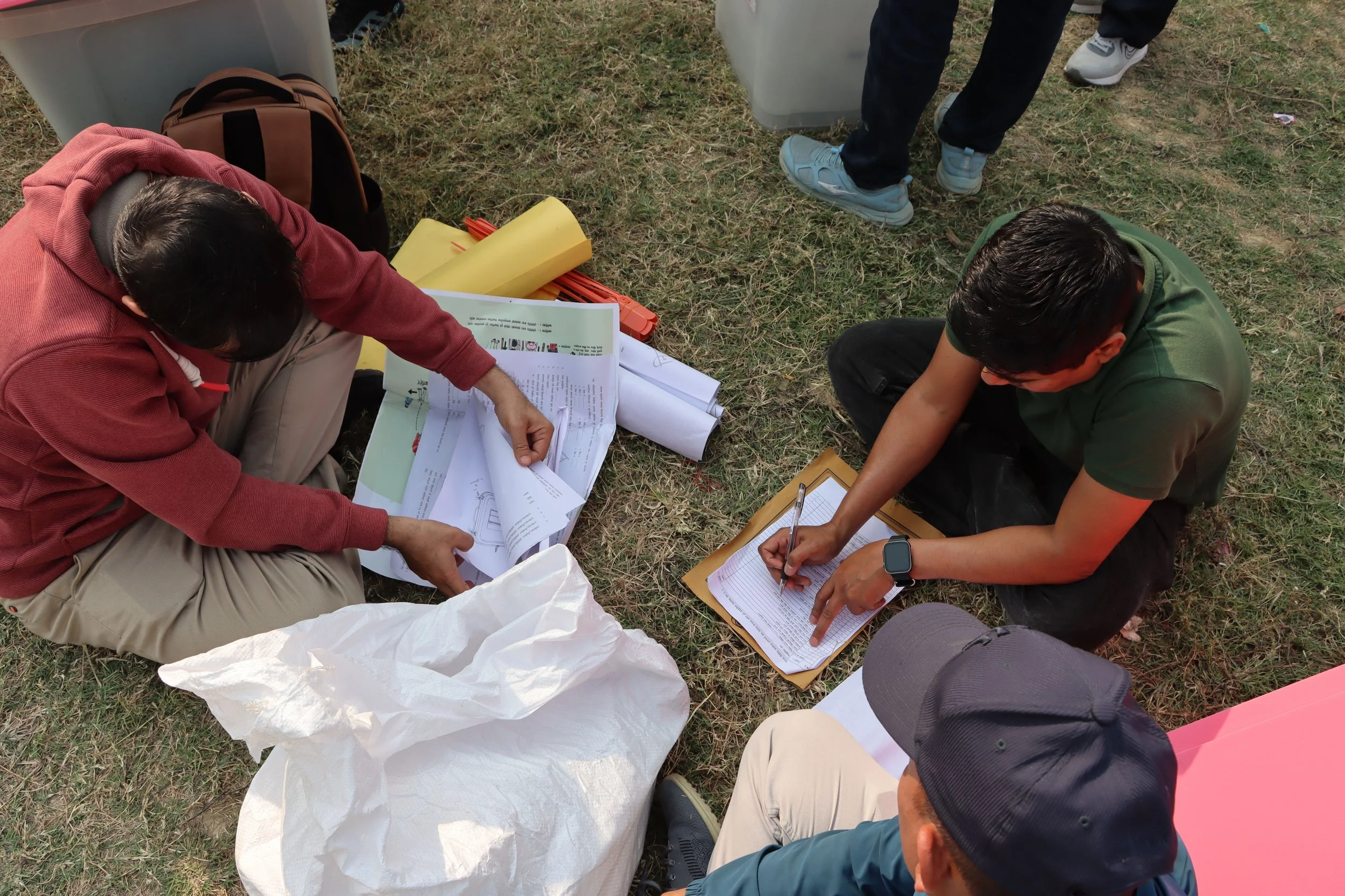 People gathered outdoors on grass, reviewing maps and documents, some sitting while others stand around, with supplies and bags nearby.