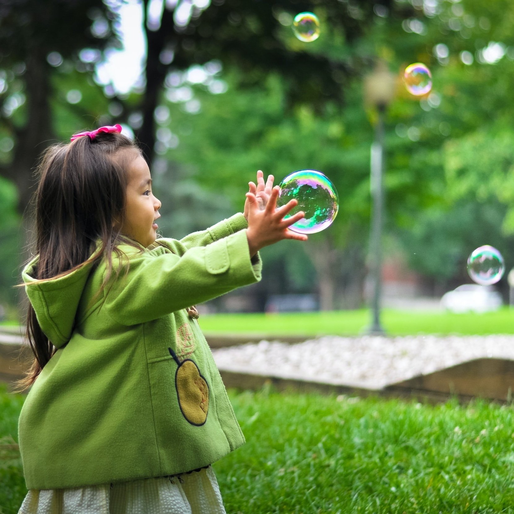 A young girl in a green hoodie with a yellow cartoon character, reaching out to touch floating bubbles in a park.
