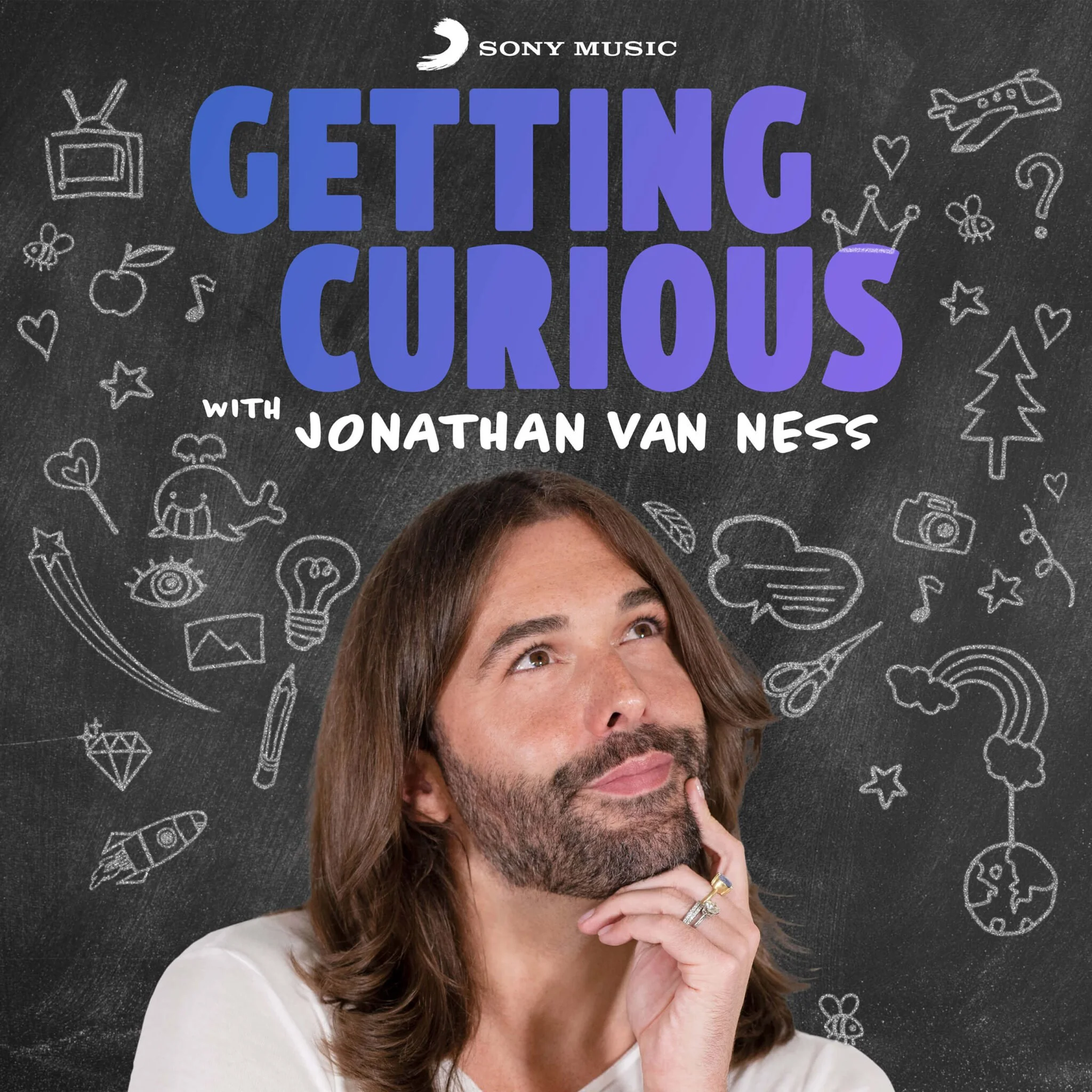 Close-up of a woman with brown hair and a beard, resting her chin on her hand, above a blackboard with white chalk drawings of various doodles, and a sign that reads 'Getting Curious with Jonathan Van Ness'.