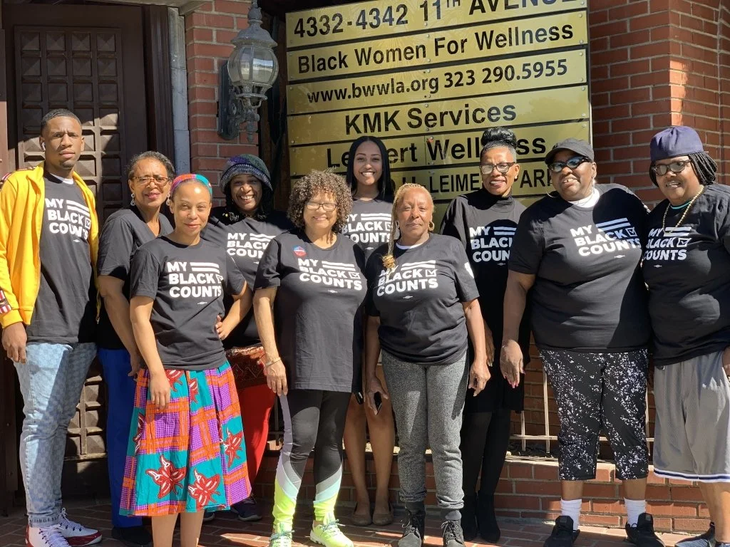 Group of diverse women standing outdoors in front of a brick building, wearing black T-shirts that say 'My Black Counts', with a yellow sign behind them listing organizations and services.