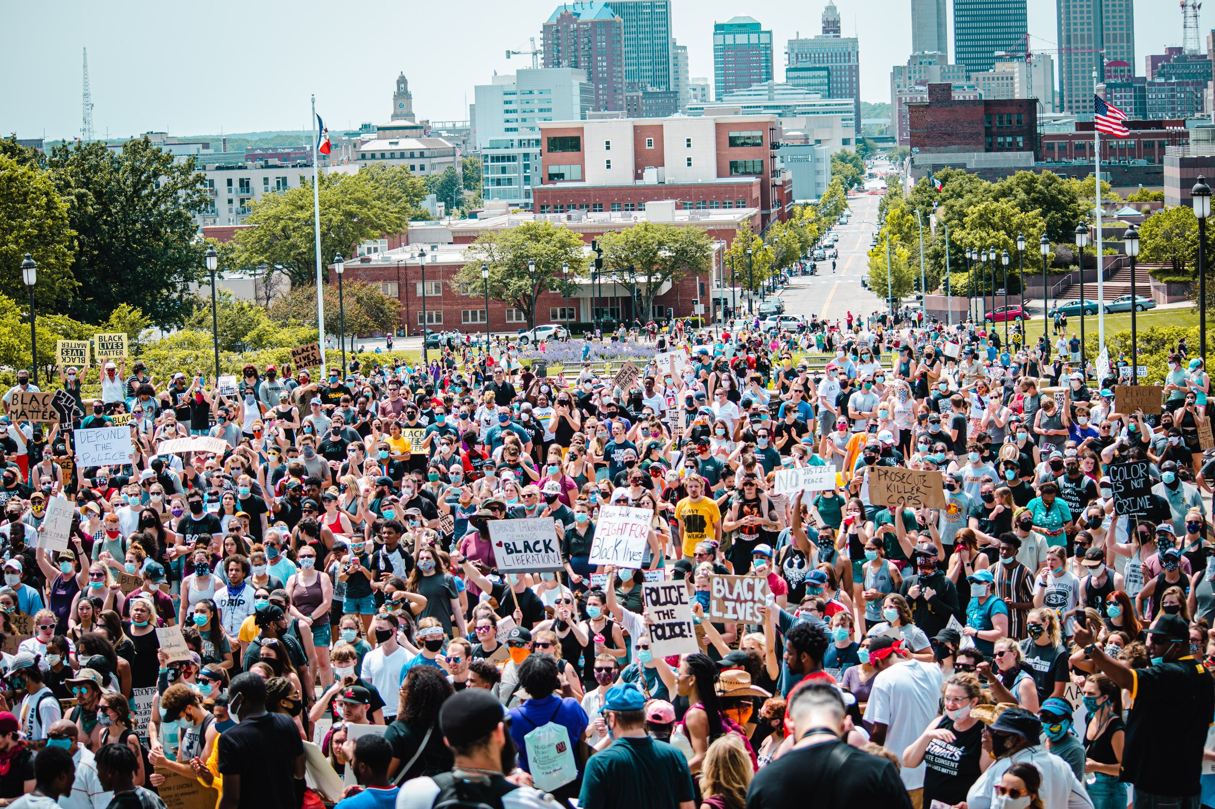 A large crowd of people gathered for a protest or demonstration in an urban setting, many holding signs with messages such as 'Black Lives Matter' and 'Defund the Police.' People are wearing face masks, and tall buildings and trees are visible in the background.