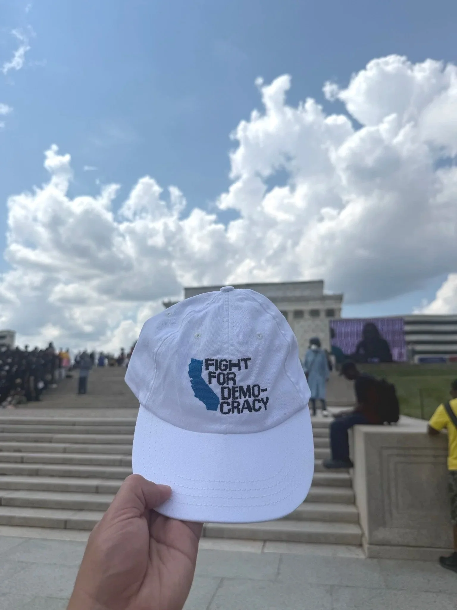Person holding a white baseball cap with the text 'Fight for Democracy' and an outline of California on it, in front of the U.S. Capitol building with steps, large crowd, and a partly cloudy sky.