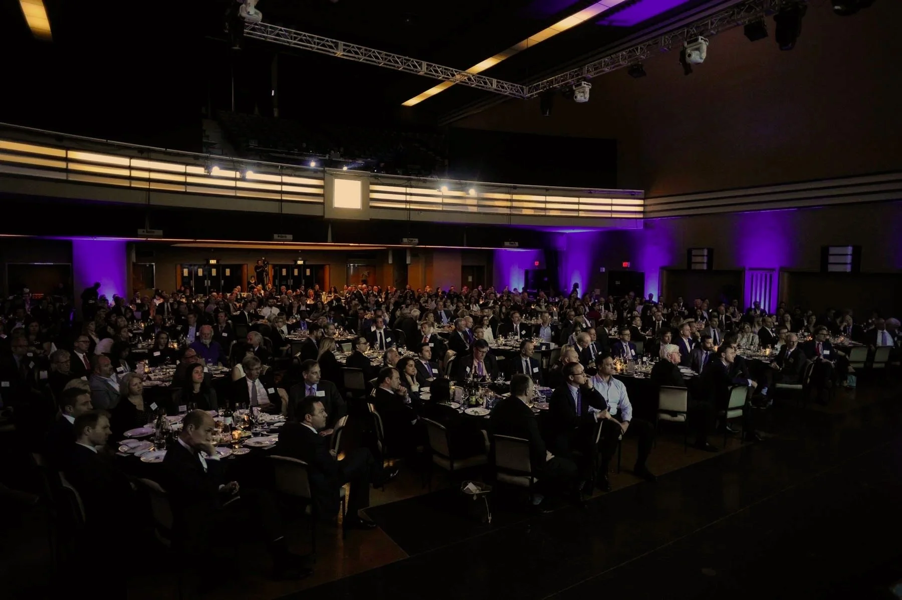 Conference or banquet hall filled with many attendees seated at dining tables, illuminated with purple lighting on the walls, with a balcony and stage area in the background.