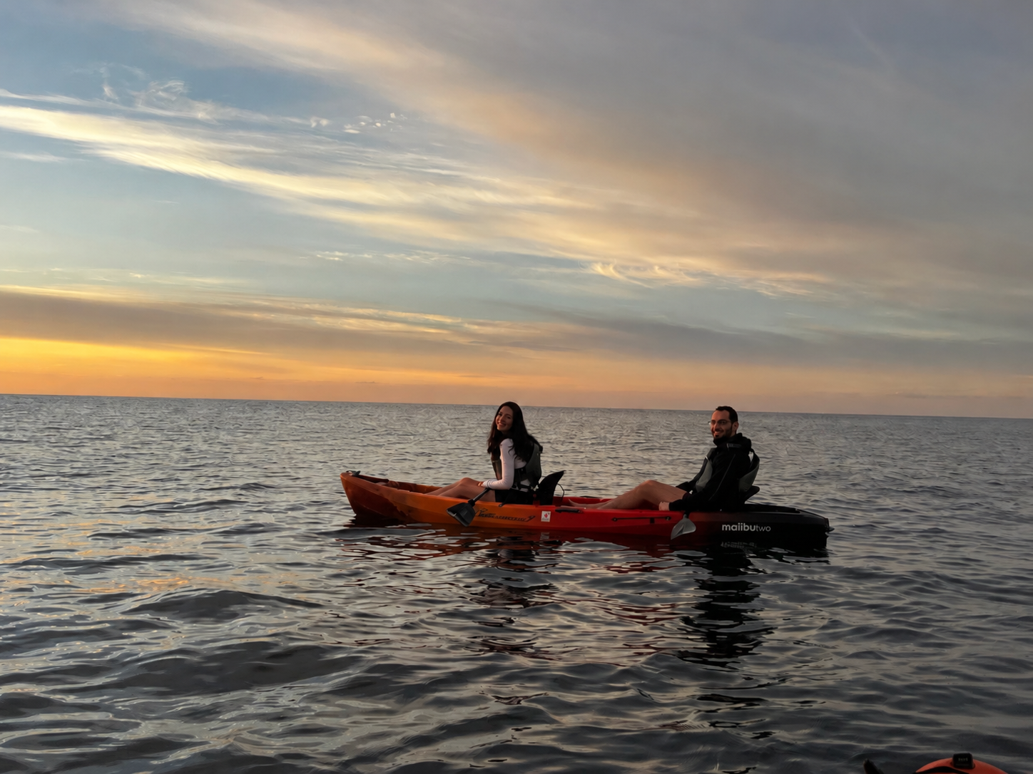 A smiling woman and man kayaking on the water during sunset, with the sky filled with colorful clouds.