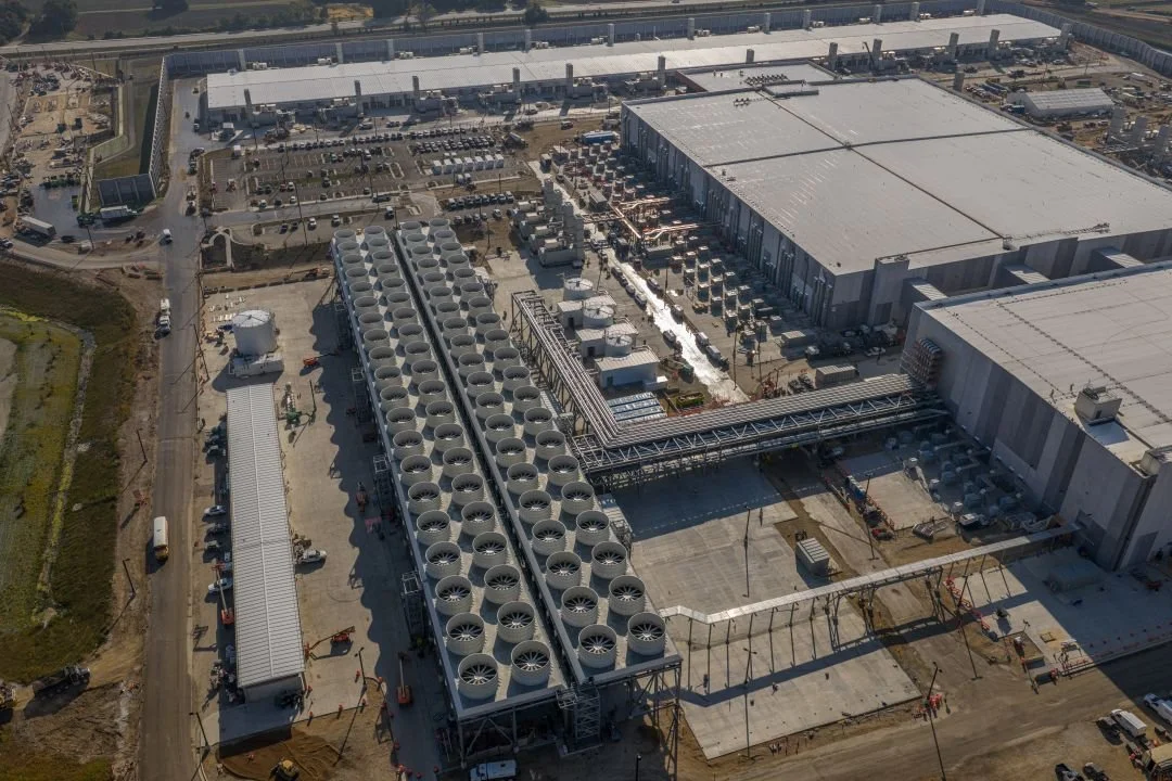 Aerial view of a large industrial facility with multiple warehouses, large fans, and a parking lot filled with vehicles.