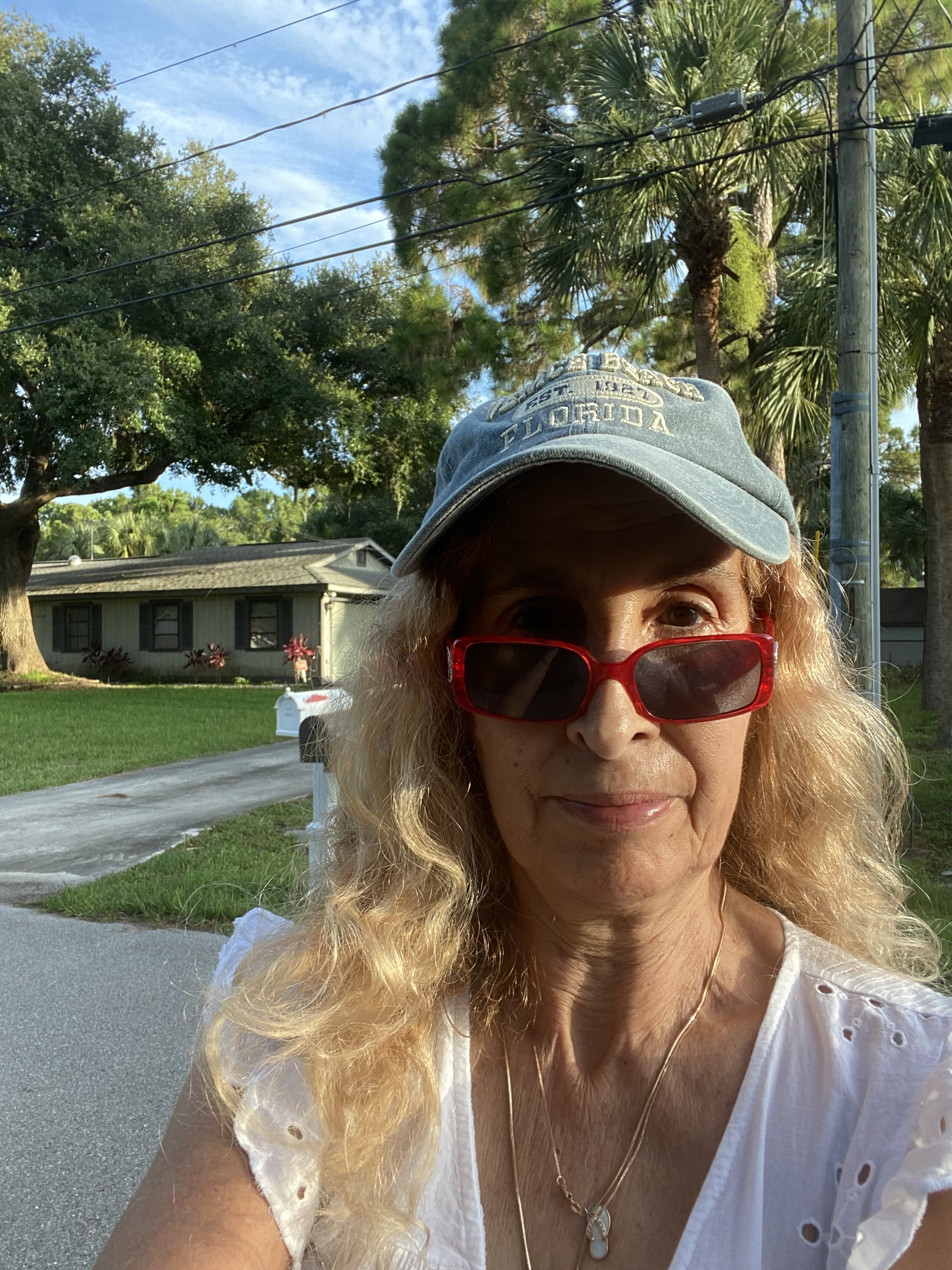 A woman with long blonde hair wearing red sunglasses, a white blouse with eyelet details, a gold necklace, and a baseball cap with 'Florida' written on it. She is outdoors near trees, a house, and a mailbox.