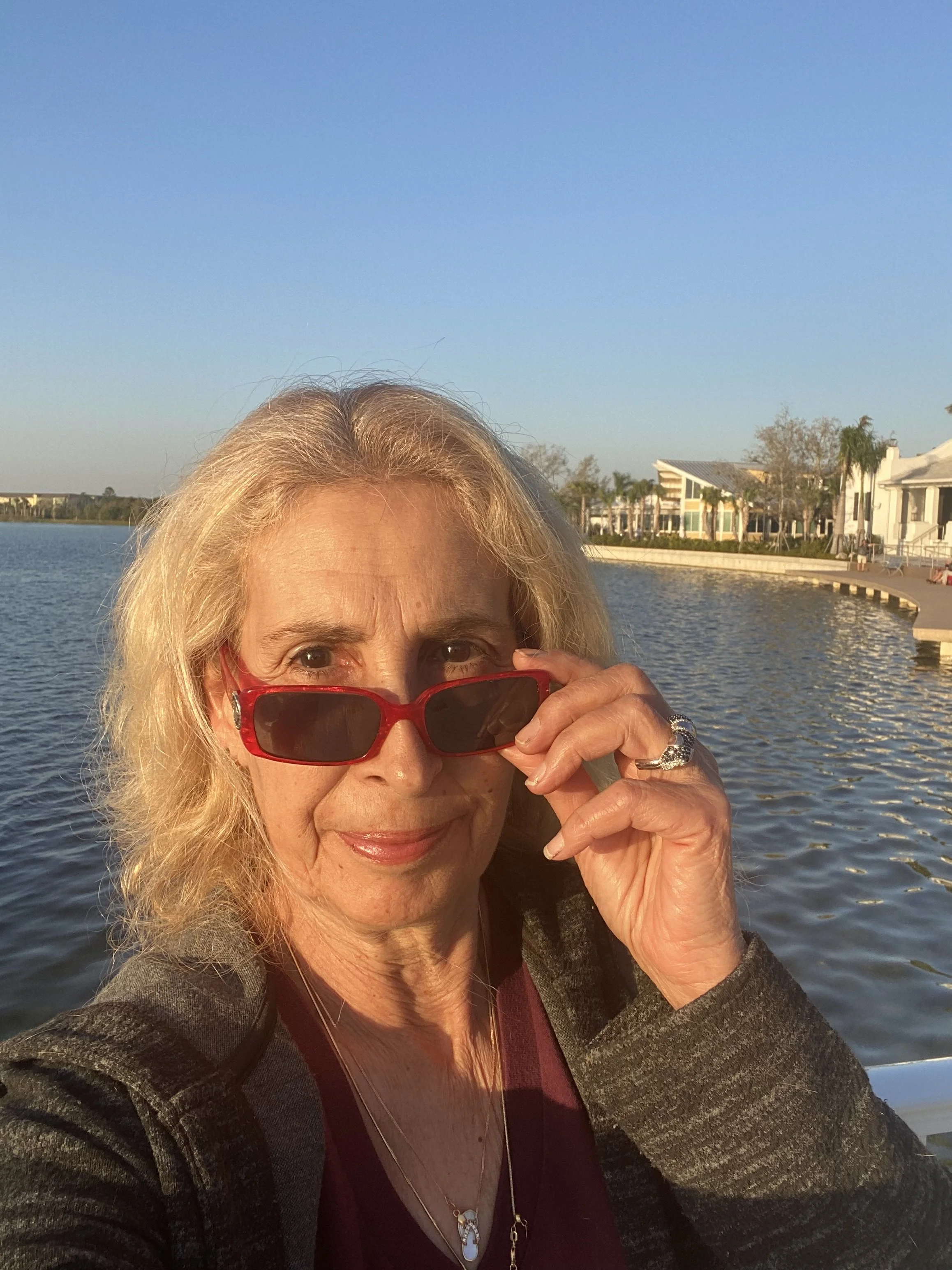 A woman with blonde hair and glasses, holding her glasses with her right hand, smiling, near a body of water with modern buildings and trees in the background during sunset.