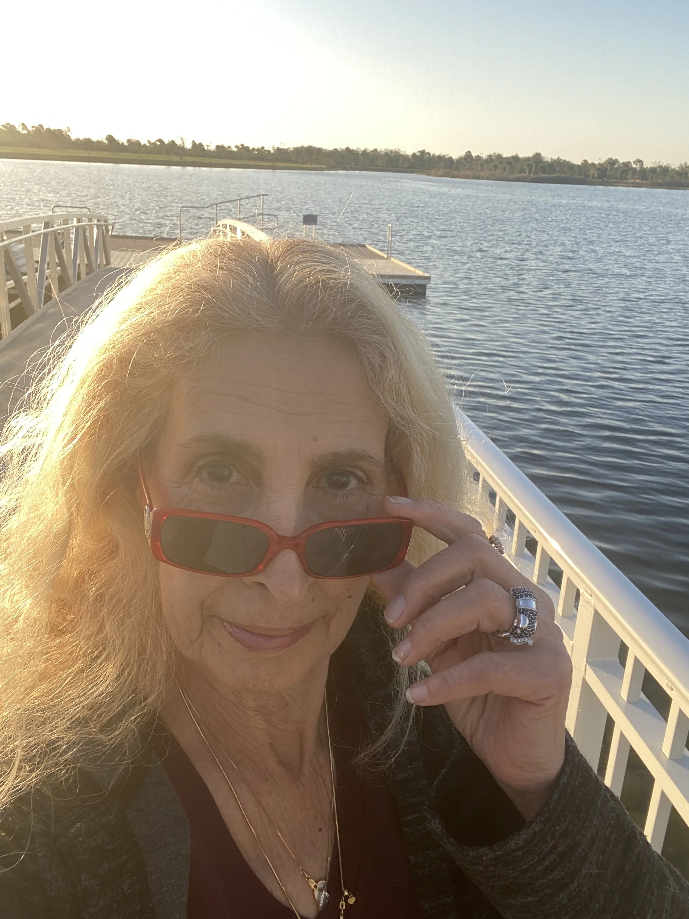 A woman with blonde hair wearing red sunglasses, a necklace, and a ring, taking a selfie on a boat by a lake during sunset.