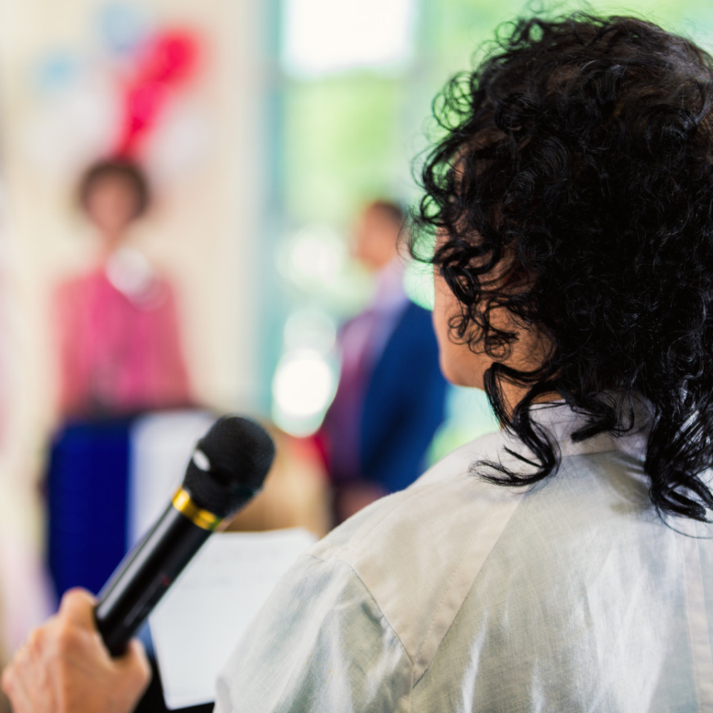 Woman with curly black hair speaking into a microphone, with children dressed in costumes or uniforms in the background.