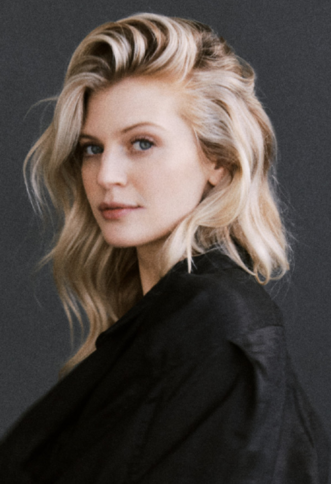 A woman with long blonde hair styled in waves, wearing a black top, posing against a dark background.