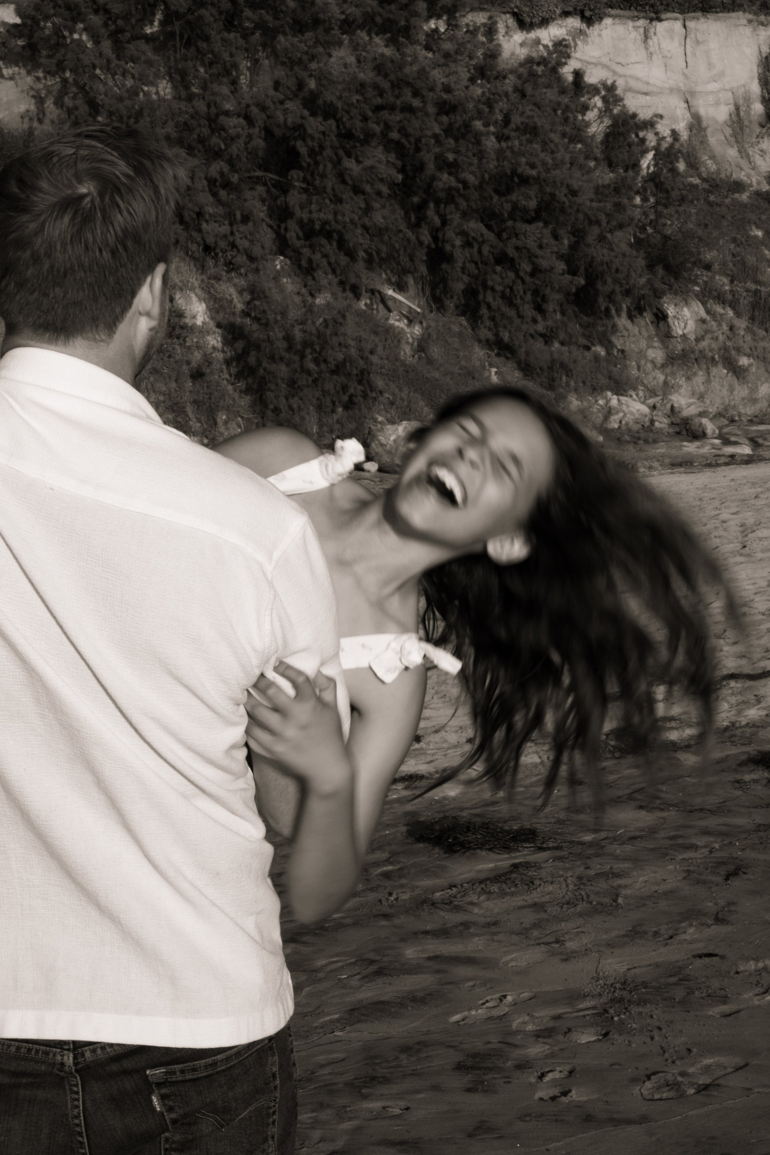 Two people, a man and a woman, standing on a beach with rocks and trees in the background. The woman appears to be laughing or smiling joyfully, while the man is facing away from the camera.