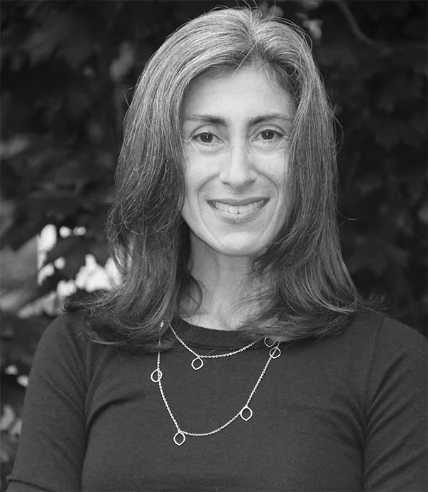 Black and white portrait of a smiling woman with medium-length hair, wearing a dark shirt and a necklace, standing outdoors.