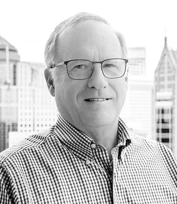A grayscale image of a man wearing glasses and a checkered shirt, standing in front of a cityscape.